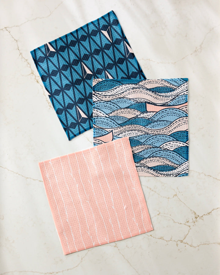 Three patterned Squall dishcloths by Twig + Spark are displayed on white marble—a super-absorbent set made from recycled materials in blue stars, blue wavy lines, and pink with vertical white stripes.