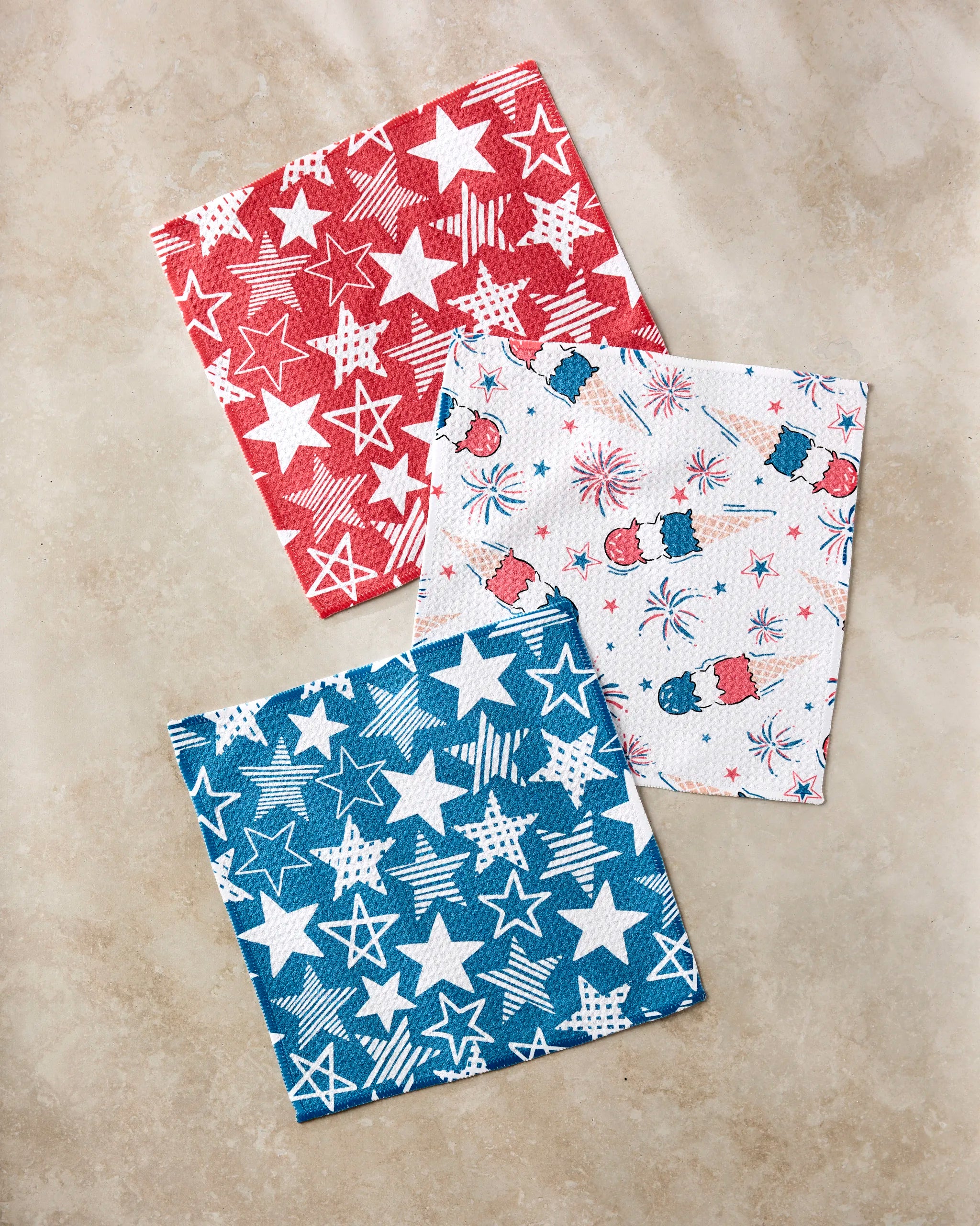 The Stars In Your Eyes cloth napkin set by Hufton Studio includes three festive recycled napkins—red with white stars, white with red and blue fireworks and hats, and blue with white stars—shown on a beige surface.