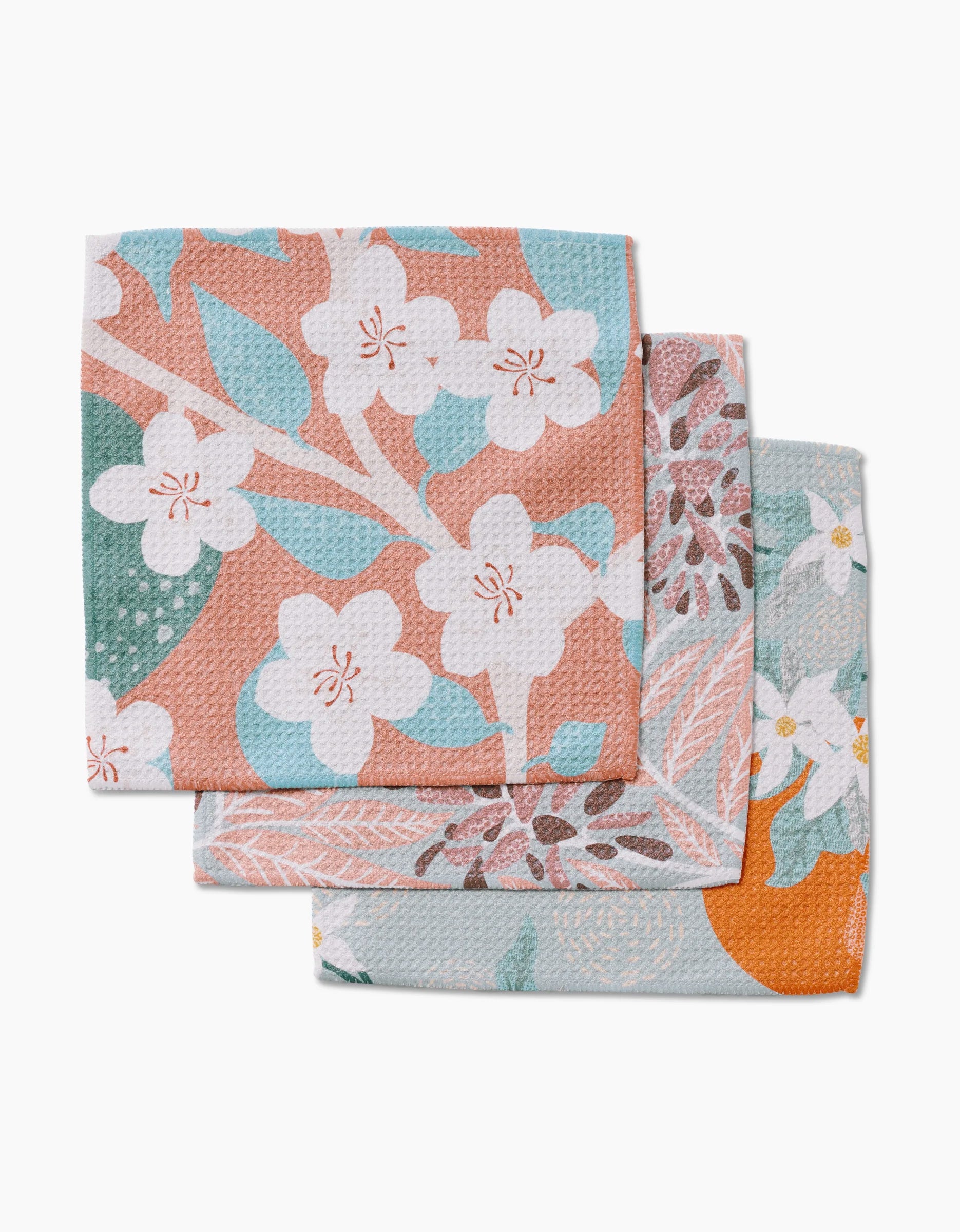 Three folded, super-absorbent State Flowers dishcloths by Julianne Haness, featuring floral and leaf patterns in peach, white, green, and orange, overlap on a white background.