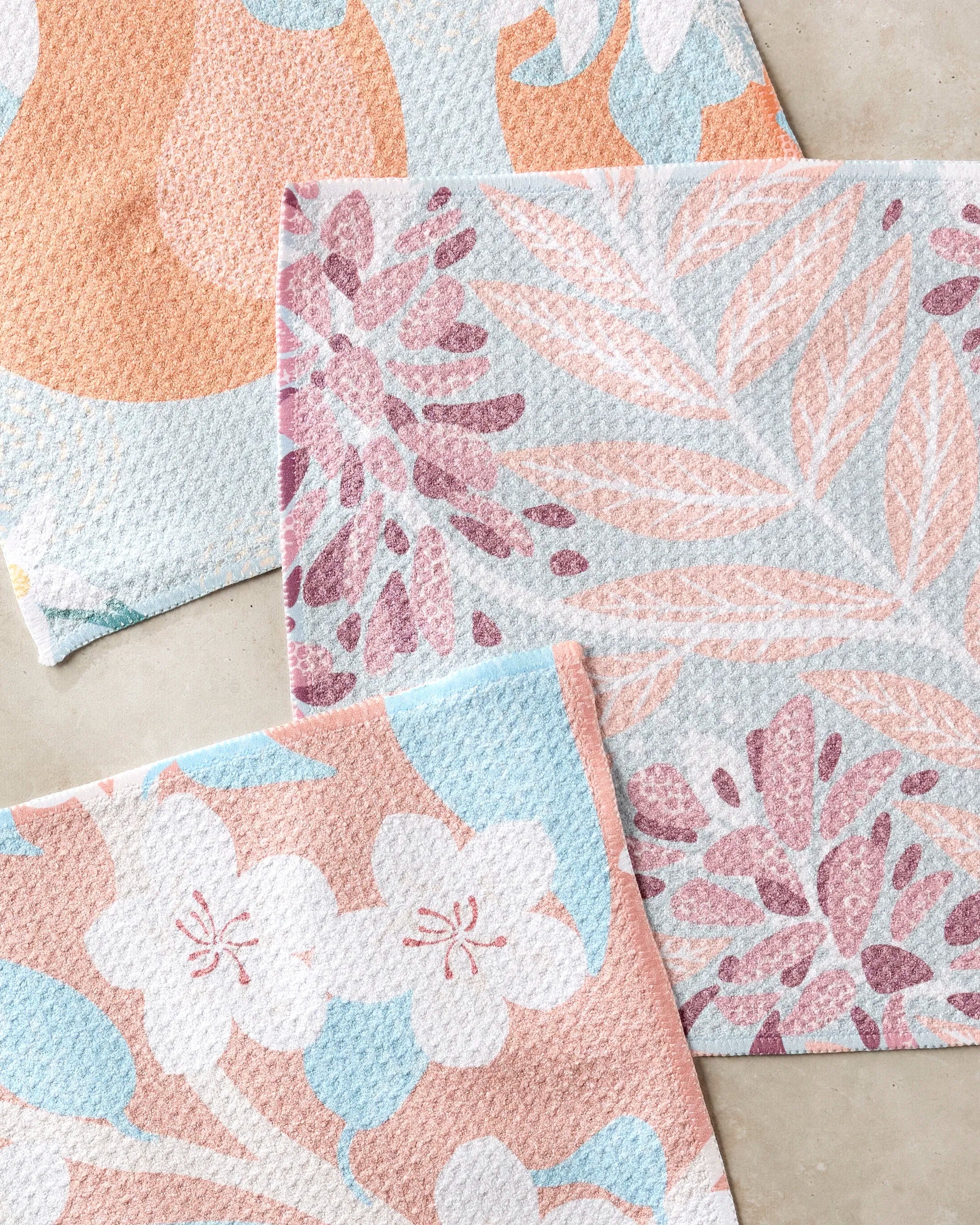 Three pastel dishcloths—State Flowers by Julianne Haness—feature floral patterns in pink, purple, blue, and white. Slightly overlapping on a beige surface, this elegant set is crafted from recycled materials.