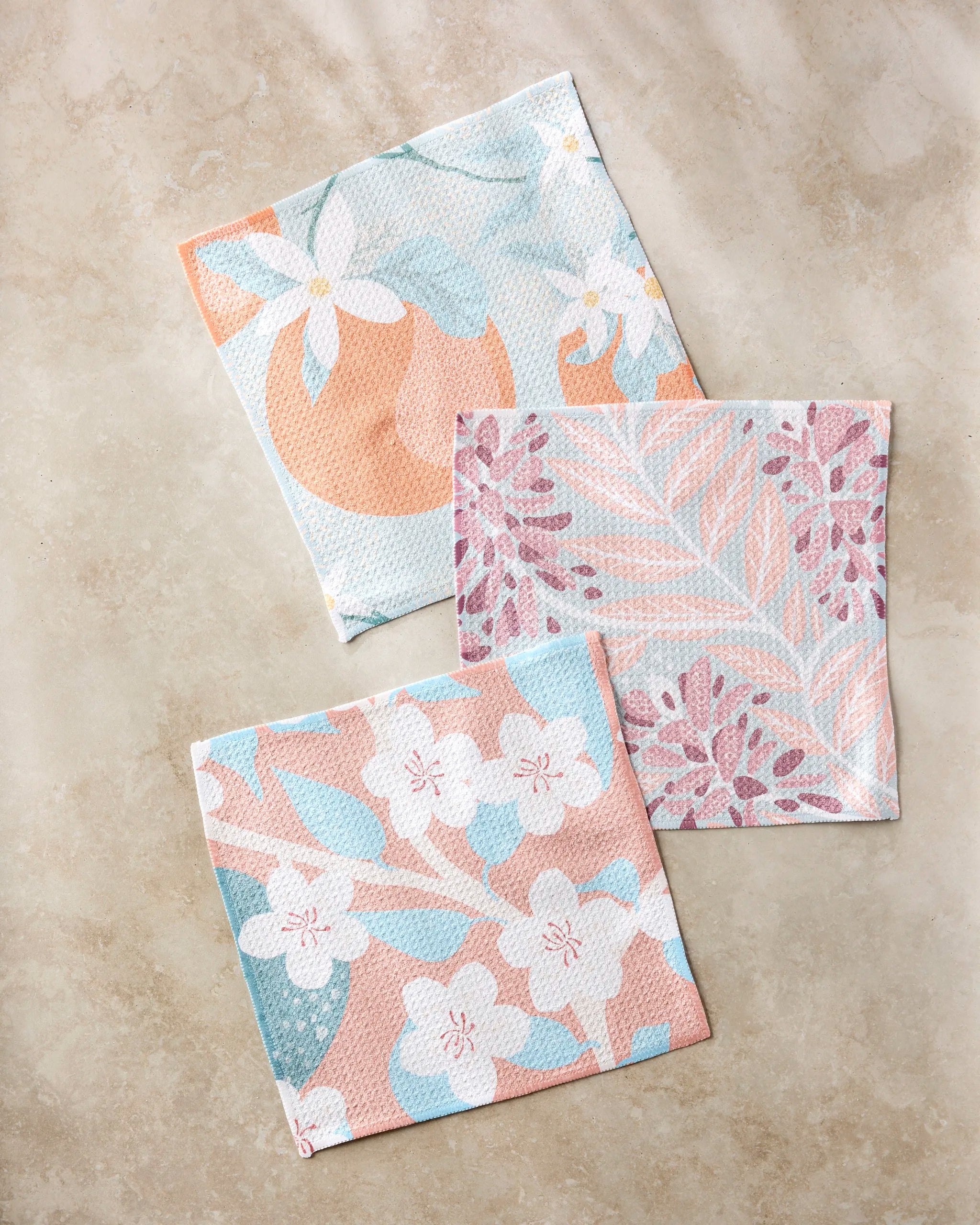 Three super-absorbent Julianne Haness "State Flowers" dishtowels with pastel floral and leaf patterns in peach, blue, and white are arranged on a light beige surface.