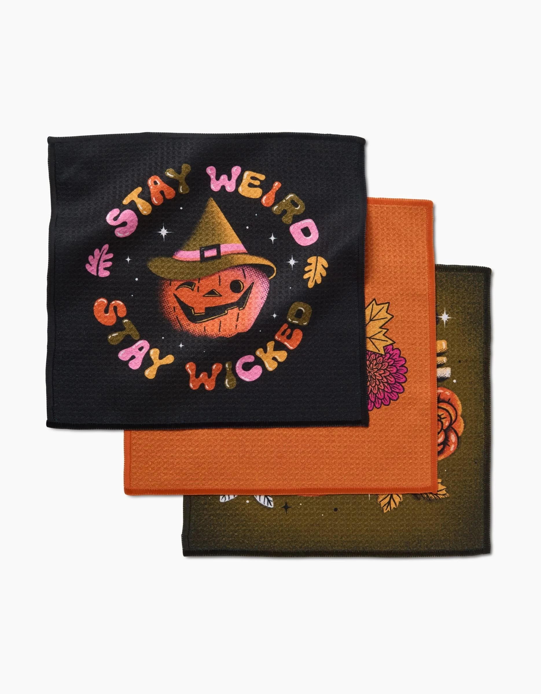 Three decorative dishcloths from Megan Roy’s “Stay Weird Stay Wicked” set: The top features a cartoon jack-o-lantern with “STAY WEIRD STAY WICKED”; the orange and dark brown pieces have floral patterns. Made from recycled materials.