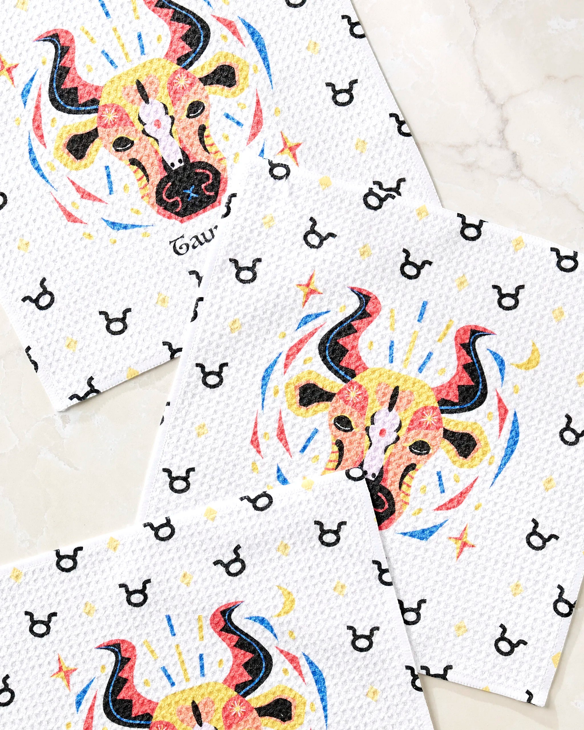 Three "Steady Taurus" napkins by Heather Zhou, featuring colorful bull illustrations and black zodiac symbols on white fabric, are displayed on marble—creating a charming dishcloth set perfect for any kitchen.
