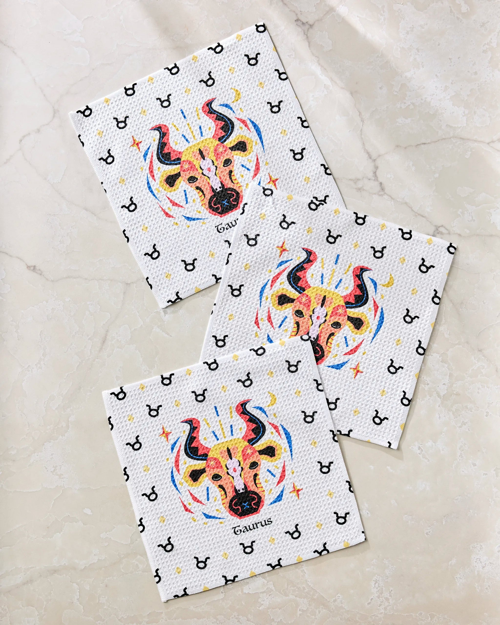 Three Steady Taurus napkins by Heather Zhou, made from recycled materials, show a colorful bull and "Taurus" with black zodiac symbols on white, displayed on light marble.