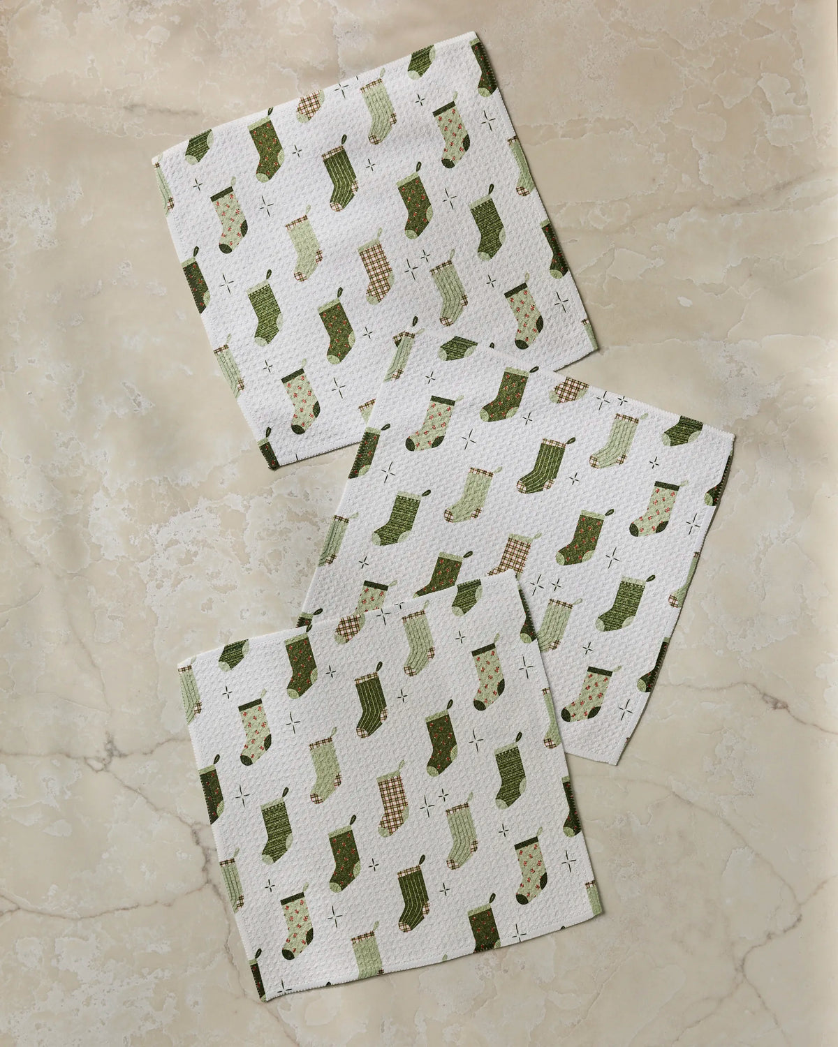 Three white absorbent kitchen towels from Geometry’s Stocking Medley collection, featuring green and brown Christmas stocking patterns, are laid out on a light beige marble surface.