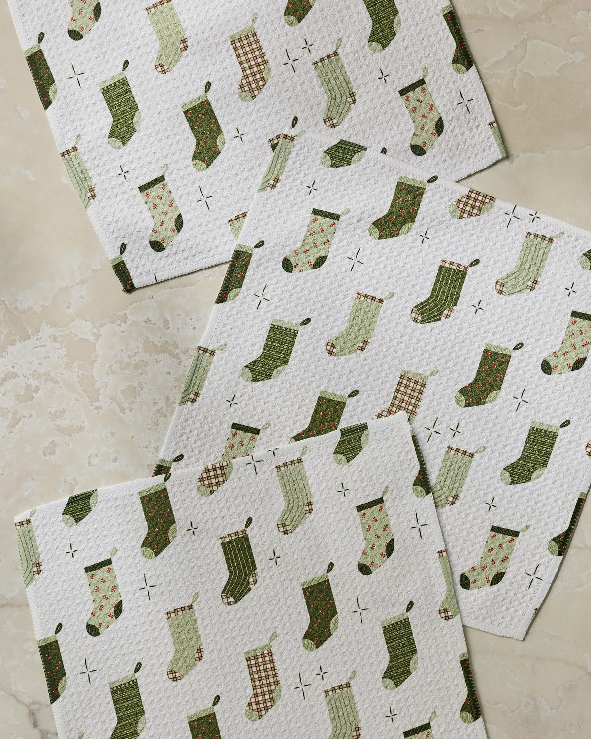Three white recycled kitchen towels from Geometry’s Stocking Medley collection, featuring absorbent fabric and festive green and plaid Christmas stocking patterns, are displayed on a light beige marbled surface. Each towel showcases a unique variation of green stockings, adding holiday charm to your kitchen décor.