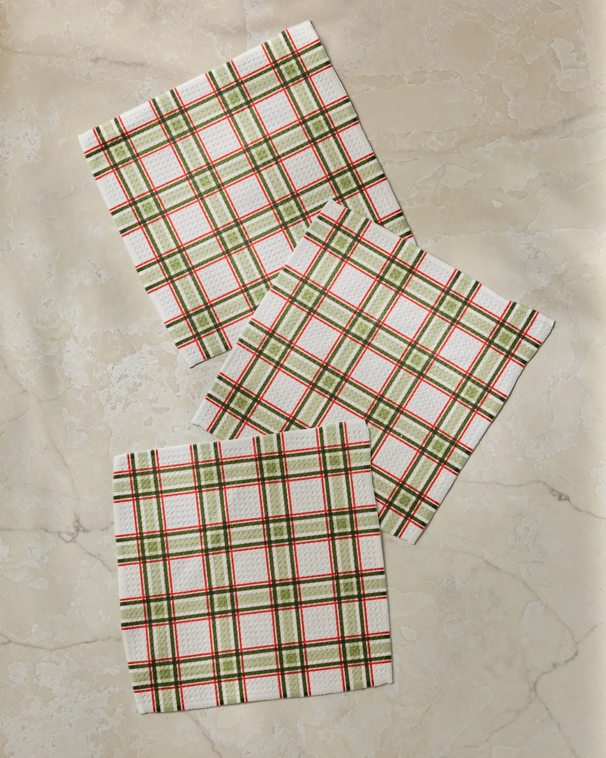Three Stocking Plaid square napkins by Geometry, made from recycled materials and featuring a red, green, and beige plaid pattern, are arranged in a loose, overlapping triangle on a light marble surface.