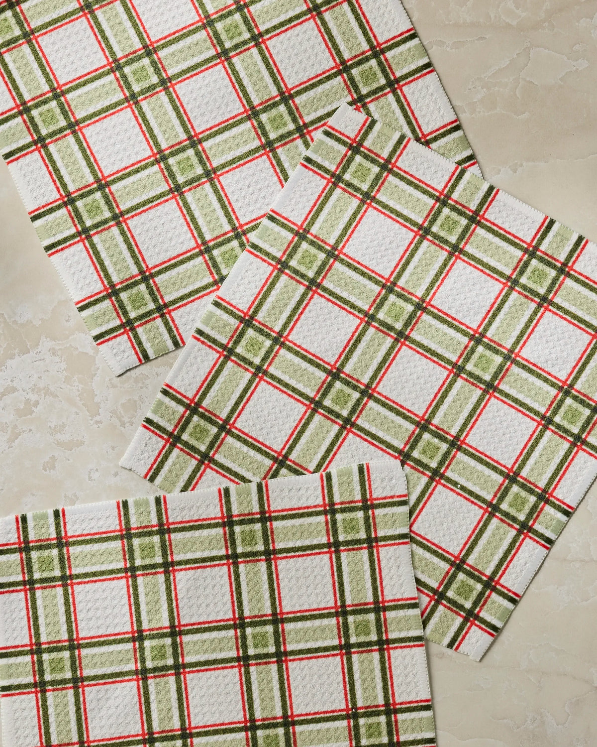 Three Stocking Plaid napkins by Geometry, featuring a classic green, red, and white plaid pattern reminiscent of a kitchen towel, are arranged on a light beige marble surface.