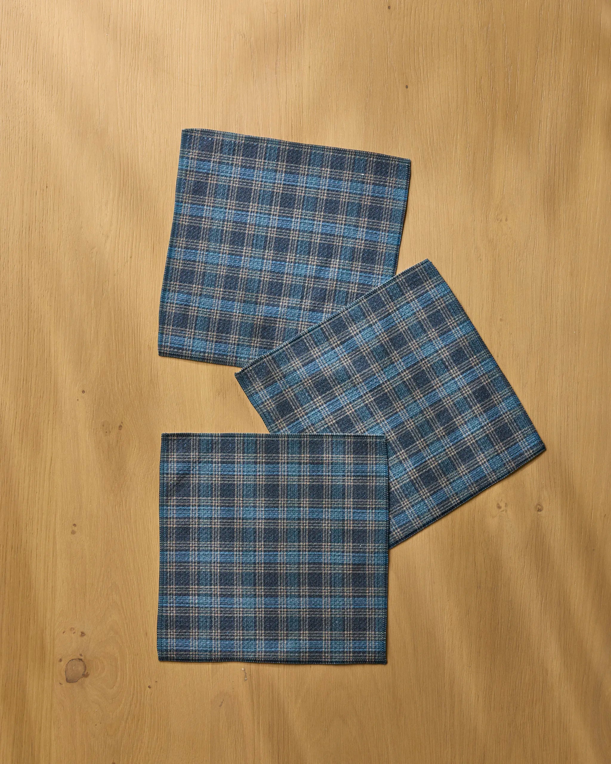 Three Stonegate napkins by Geometry, featuring a blue and gray plaid design, are arranged in a loose, staggered row on a light wooden surface—ideal as absorbent dishcloths.