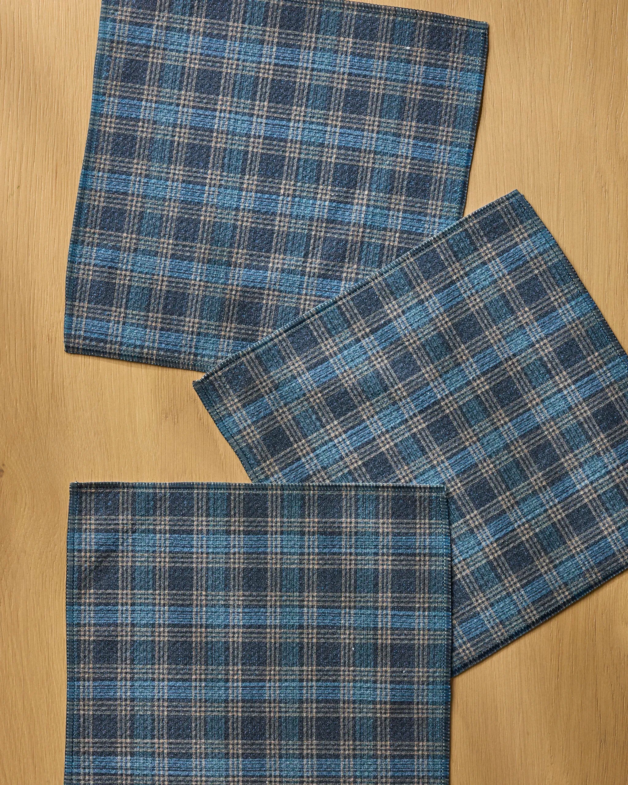 Three Stonegate kitchen towels by Geometry, featuring blue and gray plaid patterns, are neatly arranged on a light wooden surface with each towel slightly overlapping the others in a tidy geometric layout.