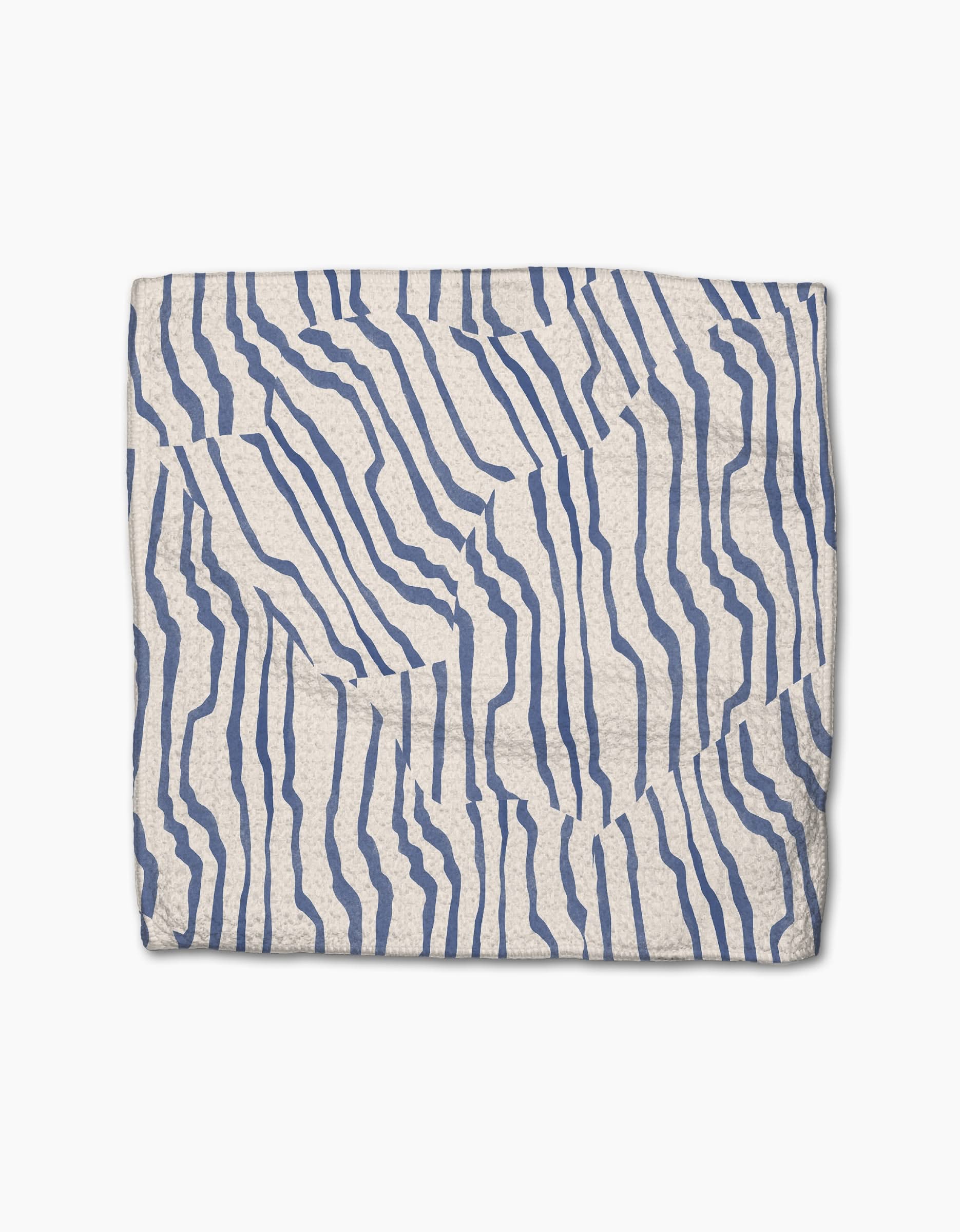 The Geometry Stripe Funk Grounds is a folded, cream-colored blanket made from recycled materials, featuring abstract wavy blue lines on a white background.
