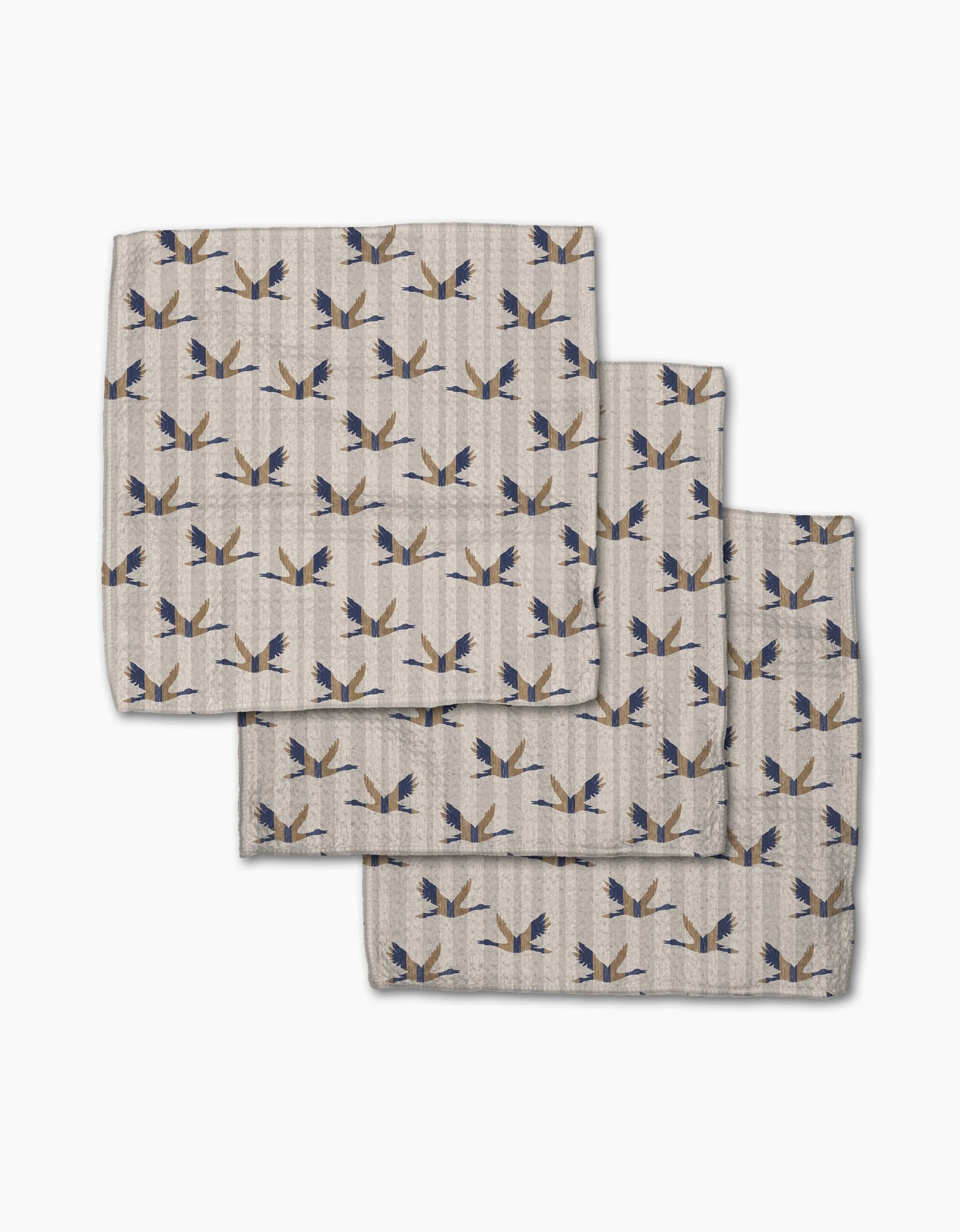 Three folded Striped Nautical Birds napkins by Geometry, featuring a blue and gold bird pattern on beige Geoweave fabric, are overlapped on a white background.