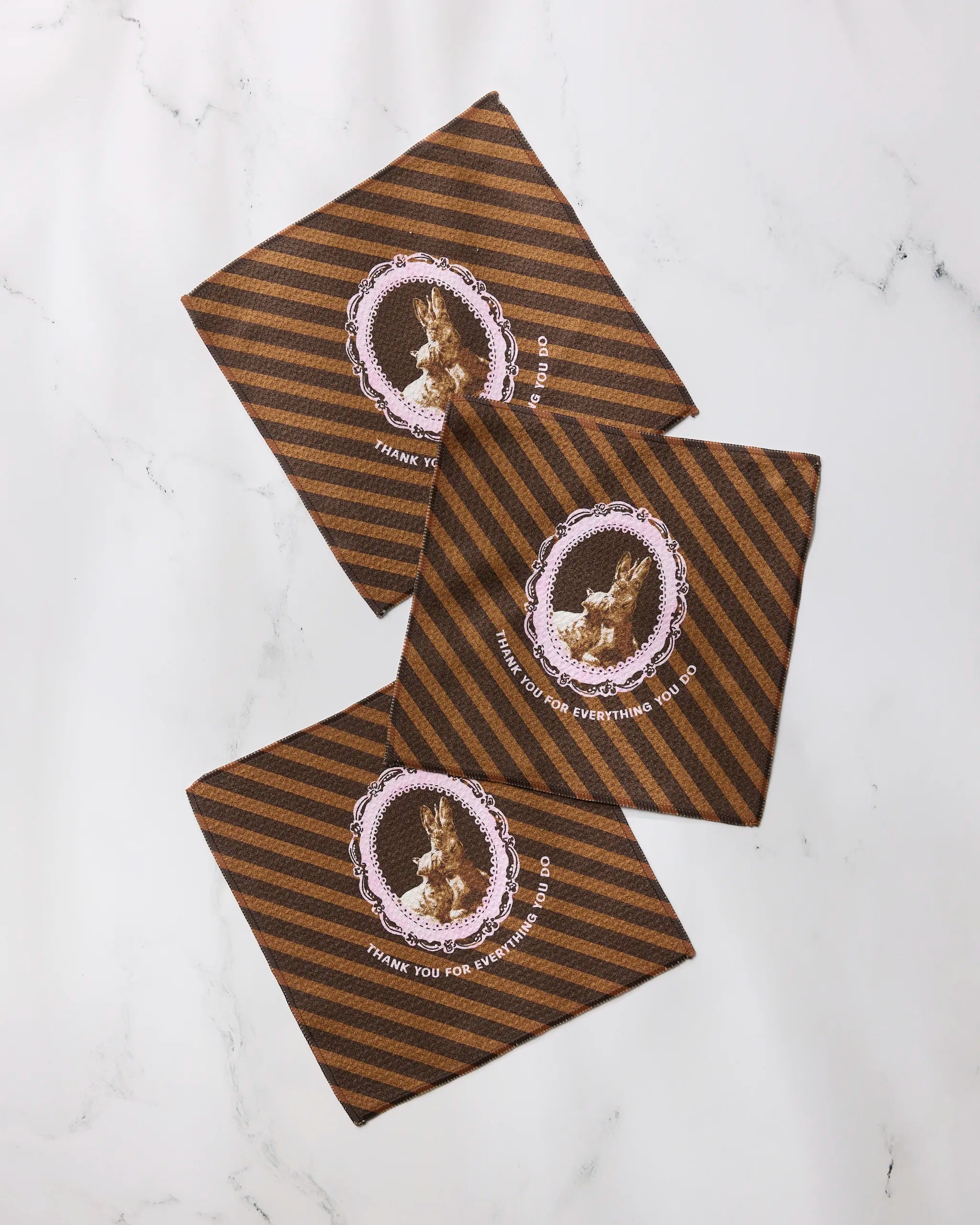 Three “Strong and Caring” napkins by Geometry, featuring brown and black stripes and decorative rabbit frames with “Thank you for everything you do,” made from recycled materials, are arranged on a white marble surface.