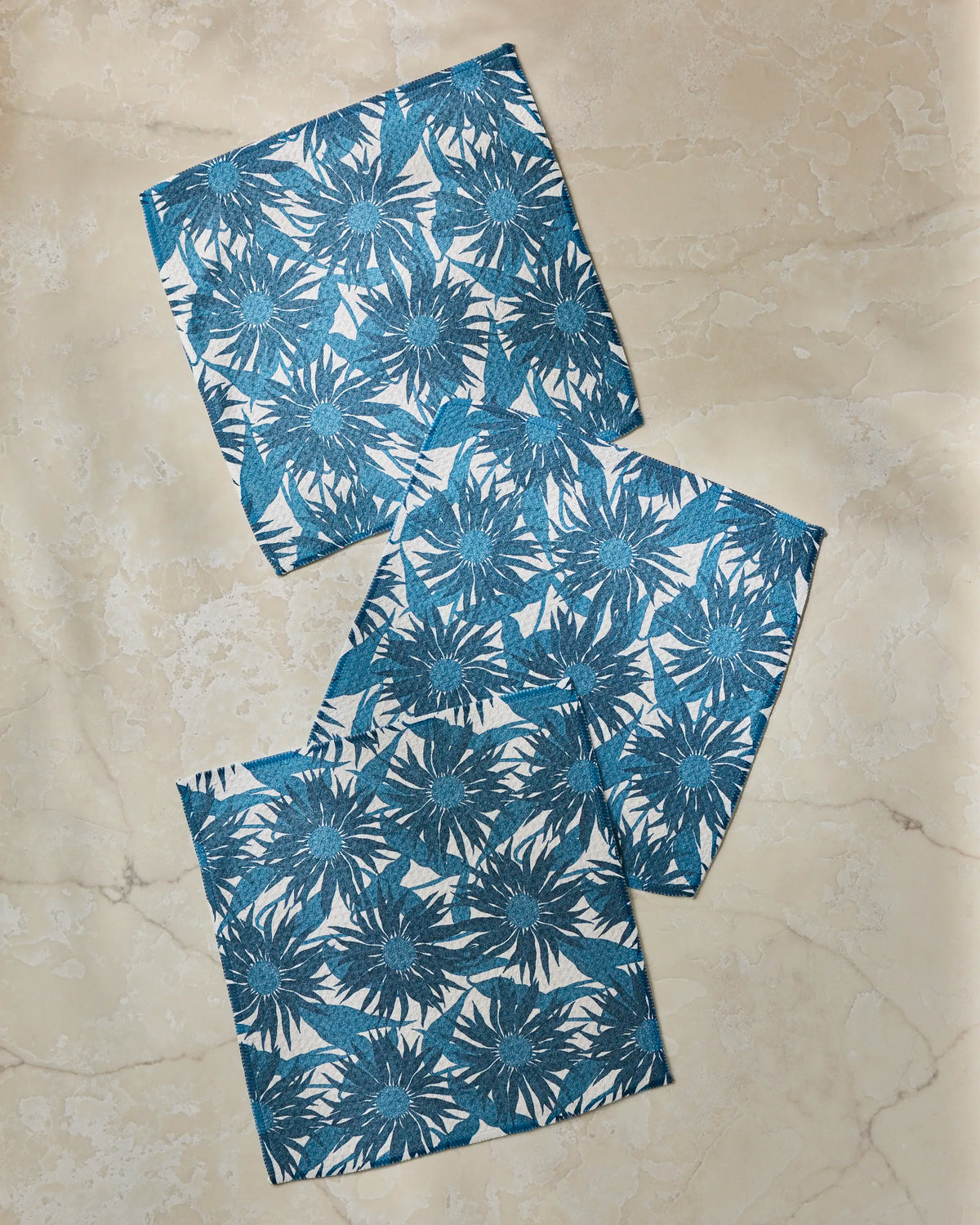 Three Sundara Frost blue and white floral-patterned cloth napkins by Geometry, crafted from recycled materials, are arranged slightly overlapping on a beige marble surface.