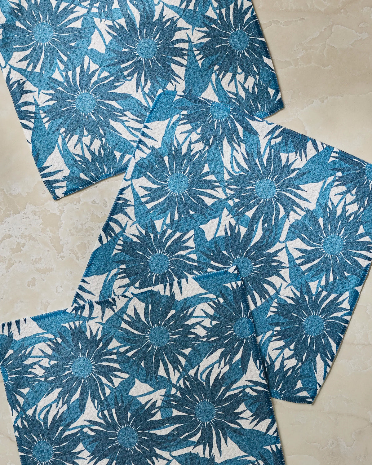 Three square Sundara Frost cloths from Geometry, featuring blue floral patterns and made with super-absorbent material, are arranged overlapping each other on a light beige marbled surface.