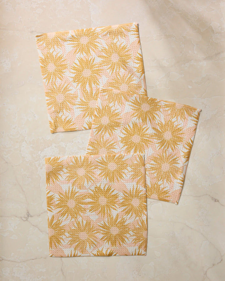 Three Sundara cloth napkins by Geometry, featuring a yellow and white sunburst floral pattern and made from recycled materials, are slightly overlapping on a light beige marble surface.