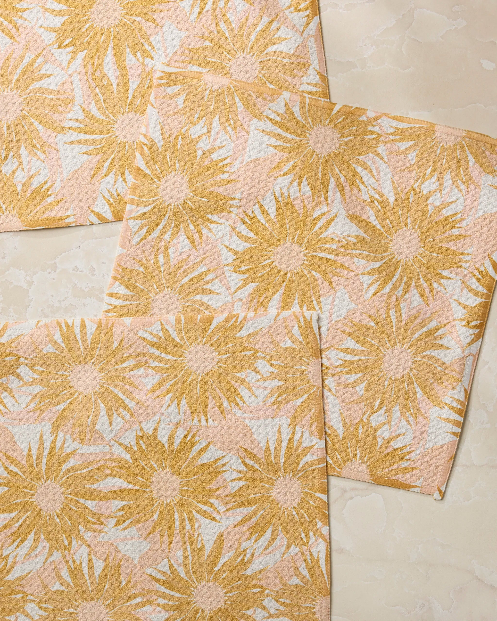 Three Sundara dishcloths from Geometry, featuring a yellow sunflower pattern on a textured beige background, are arranged overlapping on a light marble surface.