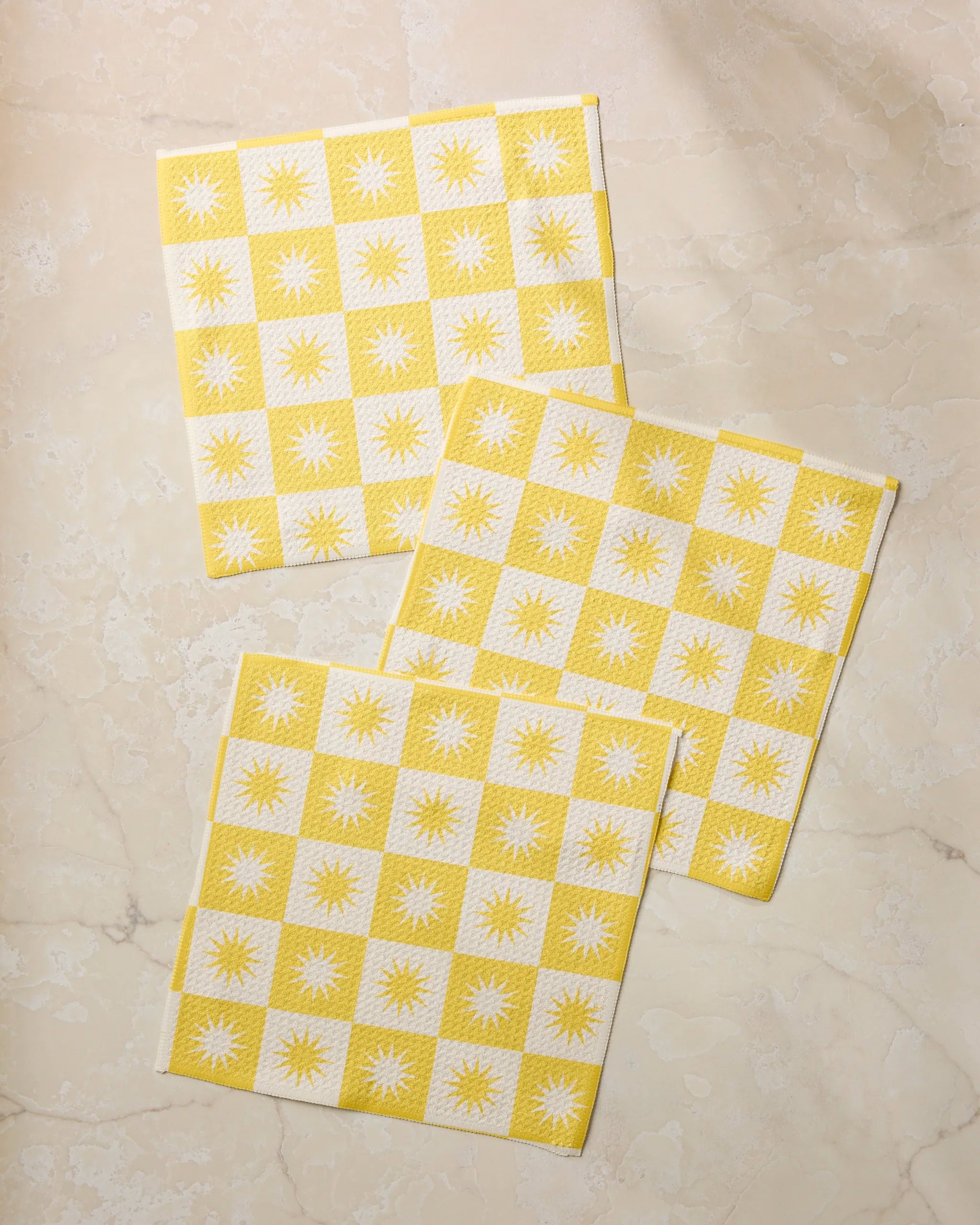 Three Sundial cloth napkins by Geometry, featuring yellow and white sunburst checkered patterns and made from recycled materials, are arranged in an overlapping diagonal layout on a light beige marble surface.