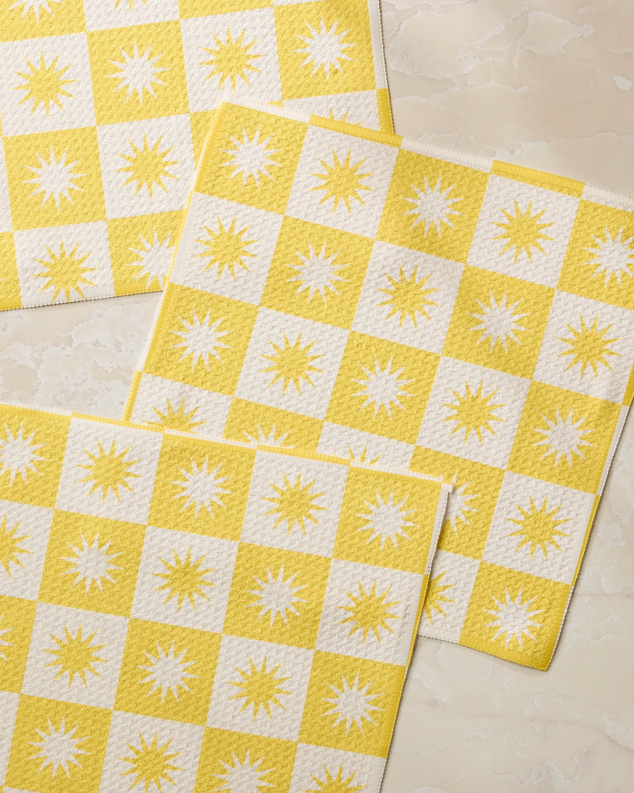 Three Sundial placemats by Geometry, featuring yellow and white checks with sunburst patterns in the white squares, made from recycled materials, are displayed on a light marble-like surface.