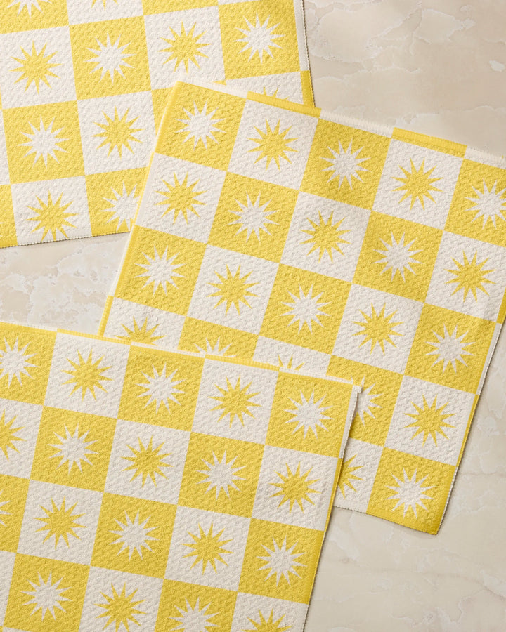 Three Sundial placemats by Geometry, featuring yellow and white checks with sunburst patterns in the white squares, made from recycled materials, are displayed on a light marble-like surface.