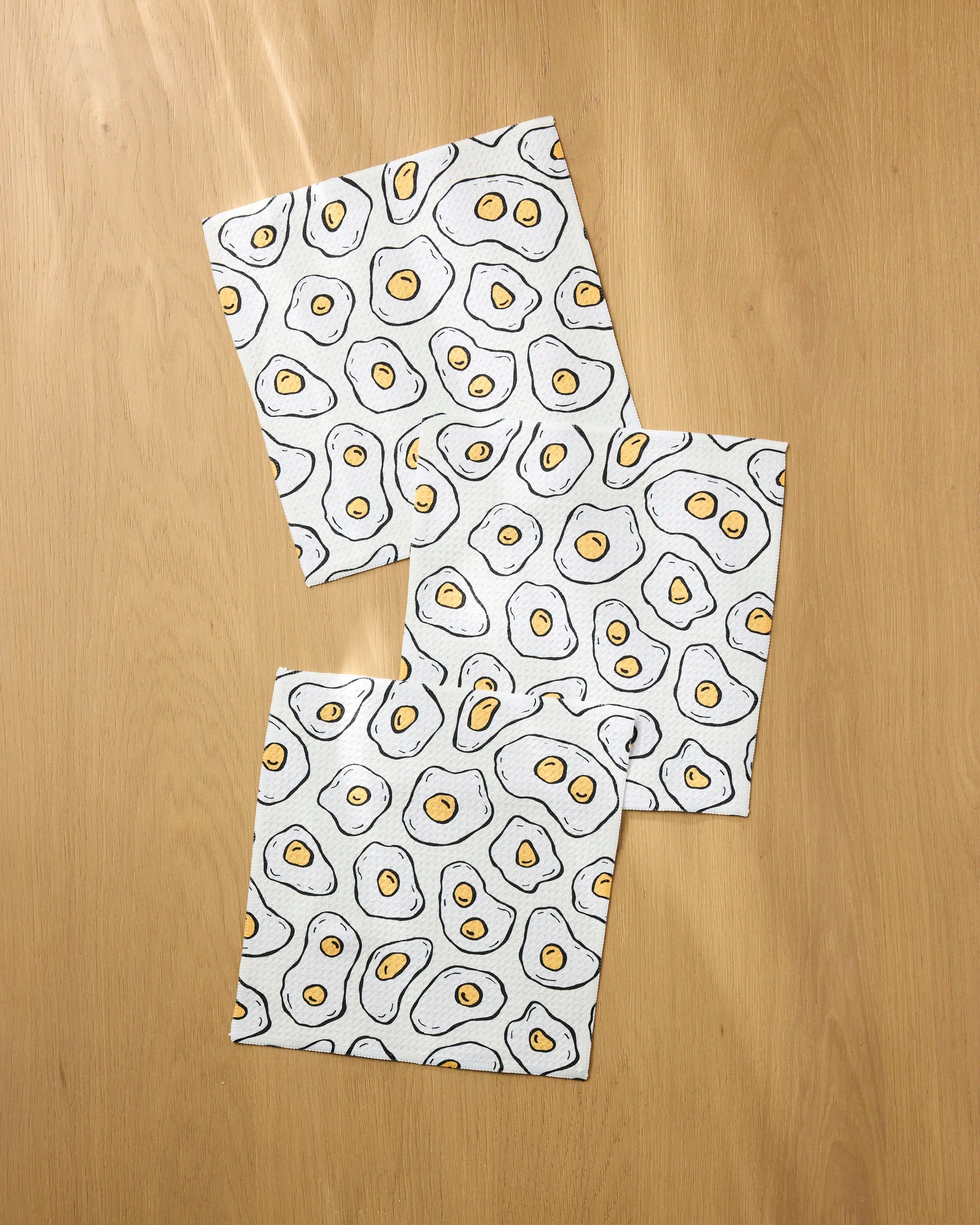 The Rosa Kichline Sunny Side Eggs set includes three eco-friendly, rectangular dishcloths with a playful fried egg cartoon pattern—perfect for adding fun and function to your kitchen.