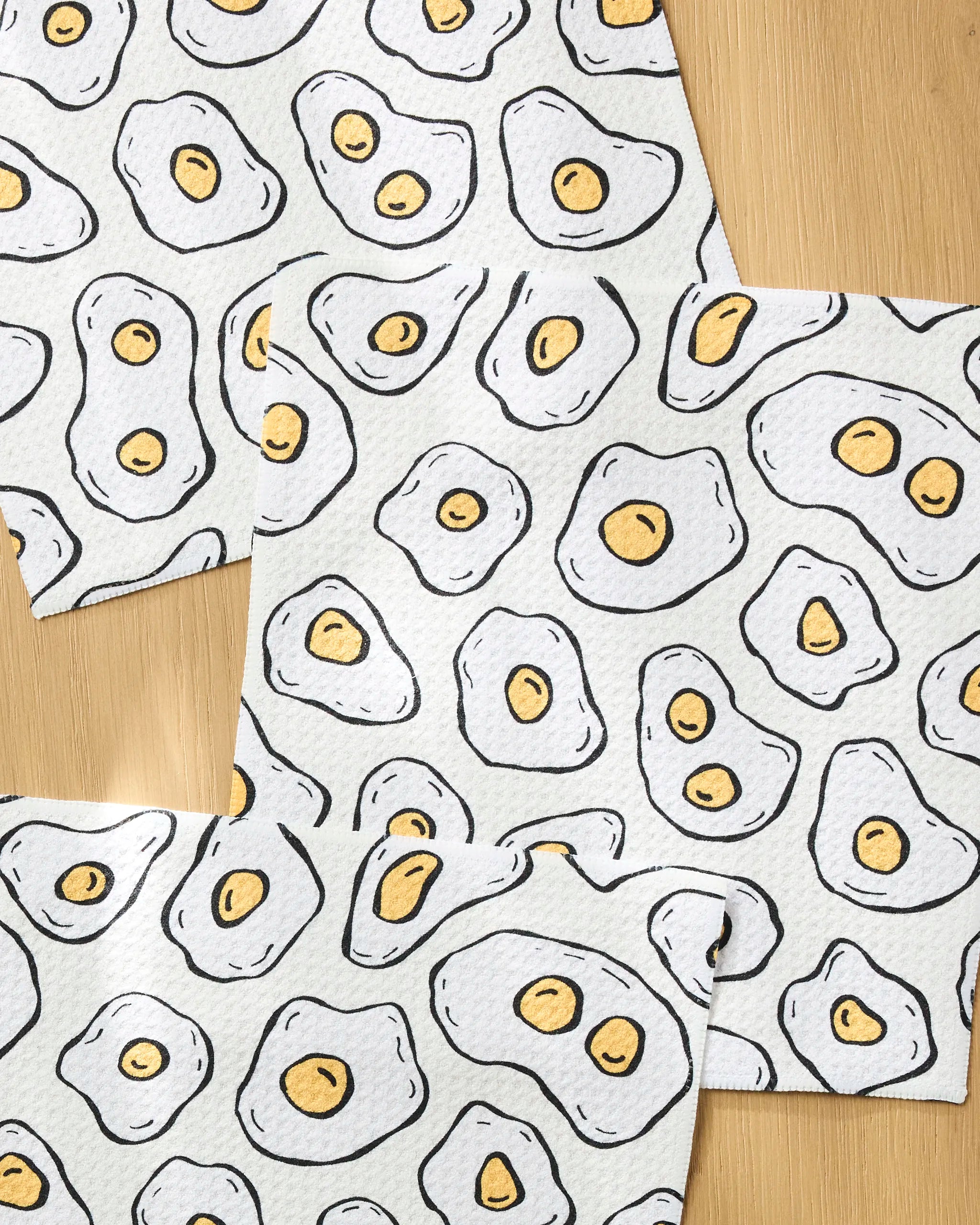 The Rosa Kichline "Sunny Side Eggs" set features three eco-friendly dishcloths with a playful pattern of cartoon fried eggs—yellow yolks and white outlines—arranged on a light wooden surface for whimsical charm.