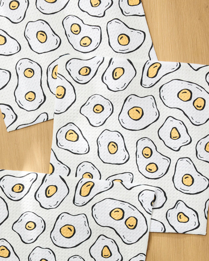 The Rosa Kichline "Sunny Side Eggs" set features three eco-friendly dishcloths with a playful pattern of cartoon fried eggs—yellow yolks and white outlines—arranged on a light wooden surface for whimsical charm.