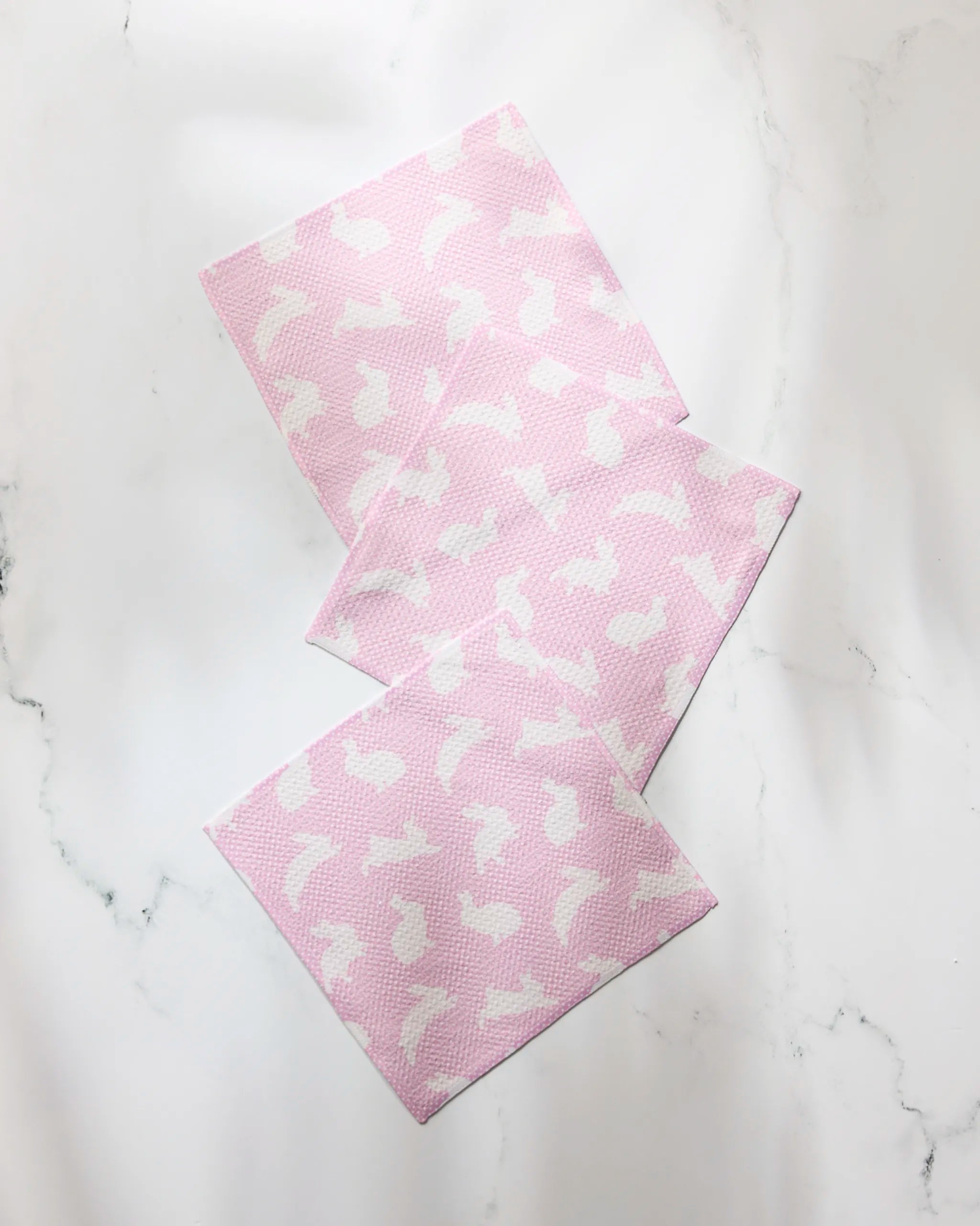 Three Geometry Sweet Bunny pink super-absorbent dishtowels, featuring a white bunny pattern, are arranged overlapping on a white marble surface.