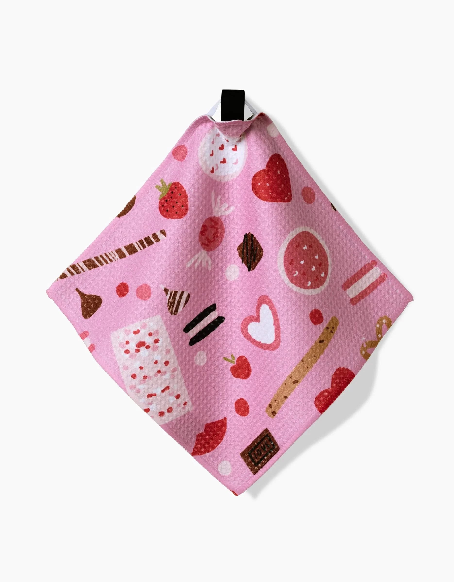 The Sweet Lovin dishcloth by Janna Sue Design is pink, textured, made from recycled materials, and features illustrations of strawberries, chocolate, cookies, hearts, and sweets. It hangs from a black loop at the top.