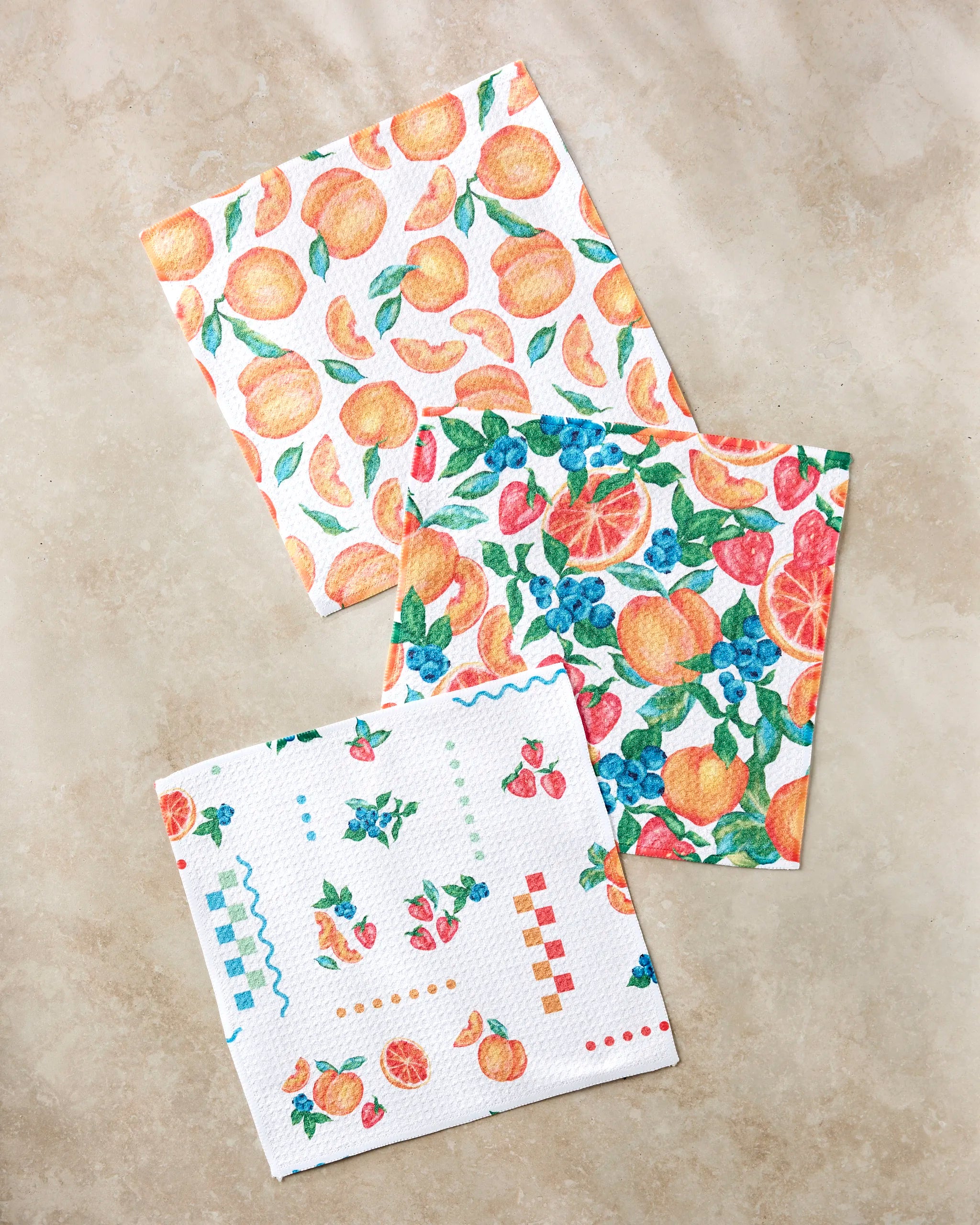 The "Sweet Pickings" set by Aubrey Fairchild features three vibrant dishcloths with fruit designs—peaches, assorted citrus and berries, plus small fruit patterns—beautifully crafted from recycled materials.