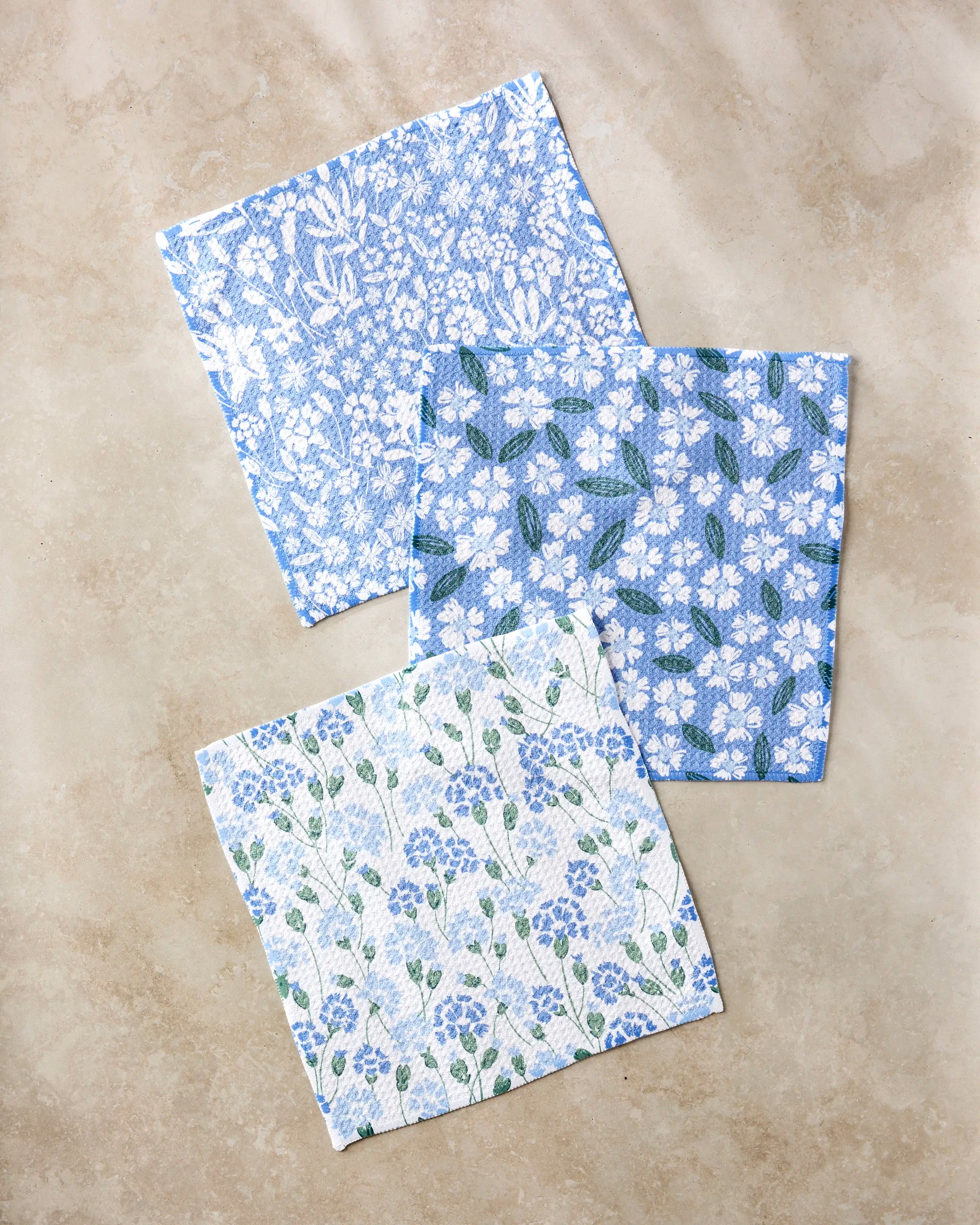 Three square Sweetie Floral dishcloths by Hufton Studio in blue, white, and green floral designs are displayed on a beige surface. Made from recycled materials, each features a unique flower pattern.