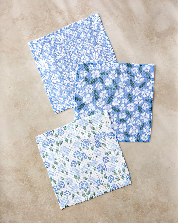 Three square Sweetie Floral dishcloths by Hufton Studio in blue, white, and green floral designs are displayed on a beige surface. Made from recycled materials, each features a unique flower pattern.