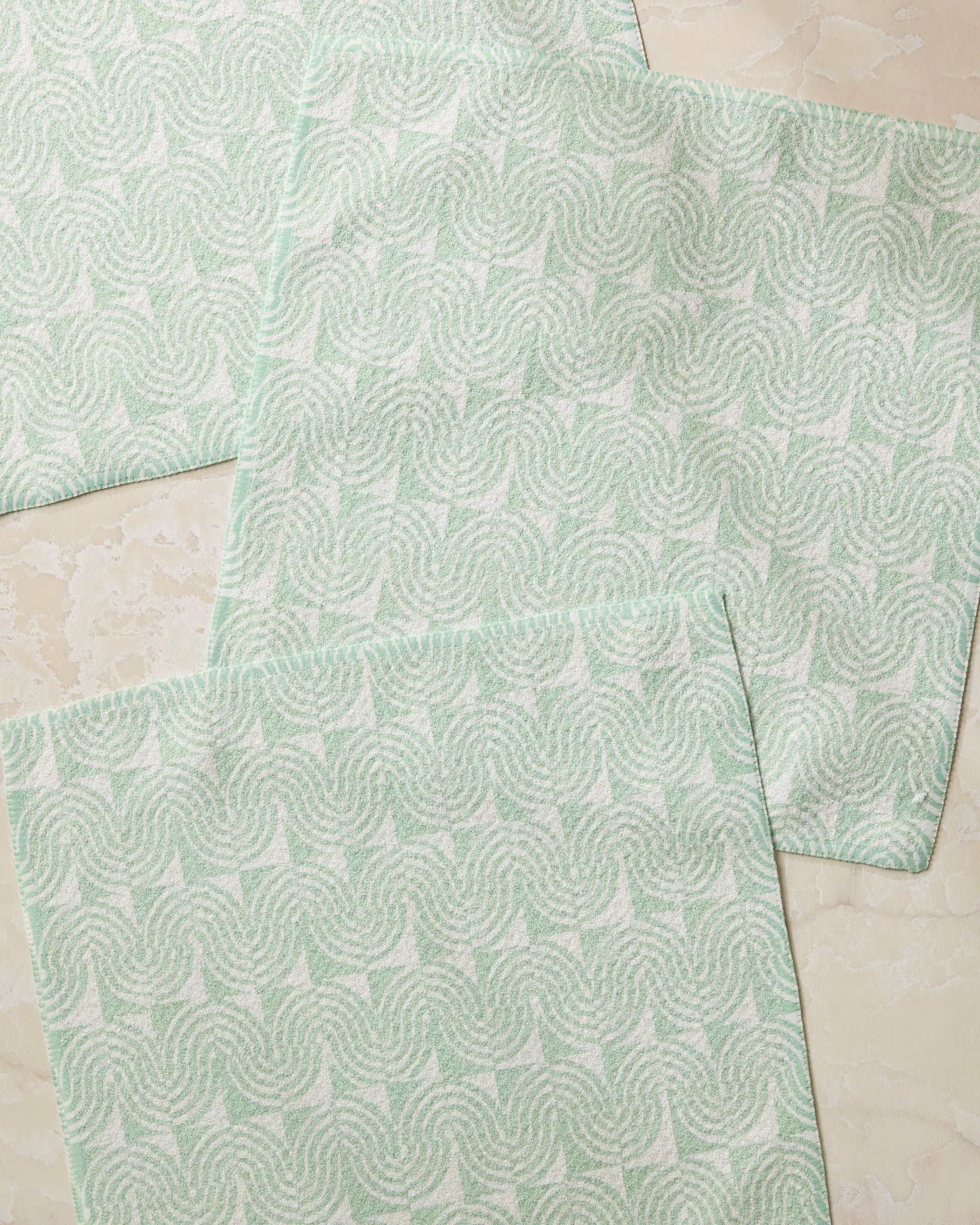 Four Swirl Land recycled dishcloths by Geometry, in pale green and white with a geometric swirl pattern, are arranged overlapping on a light marble surface.