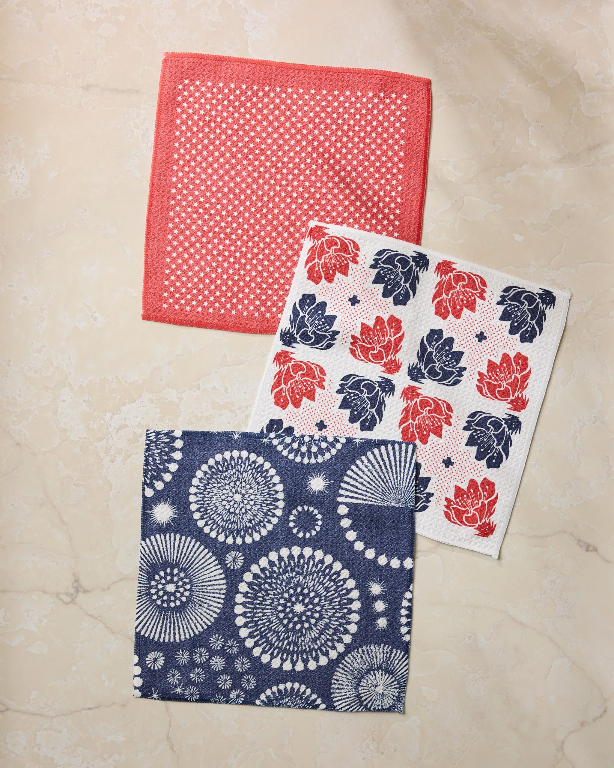Three square Talon cloth napkins by Geometry, made from recycled materials, are displayed on light marble: one red with white dots, one white with red and blue lotus flowers, and one blue with white circular floral designs.