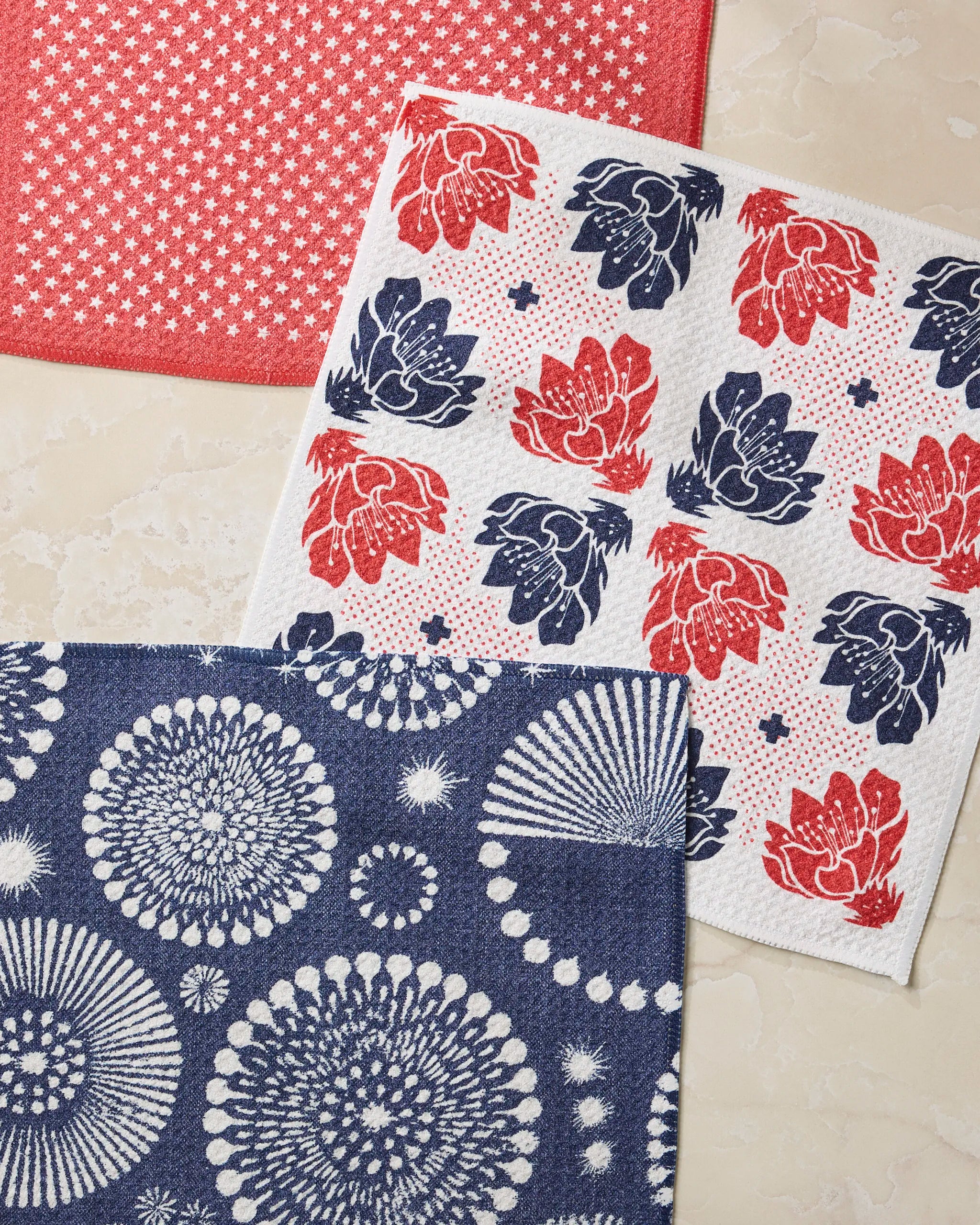 The Geometry Talon kitchen towels set includes three absorbent dishcloths: one red with white stars, one white with red and blue florals, and one navy blue with bursts, all showcased on a marble surface.