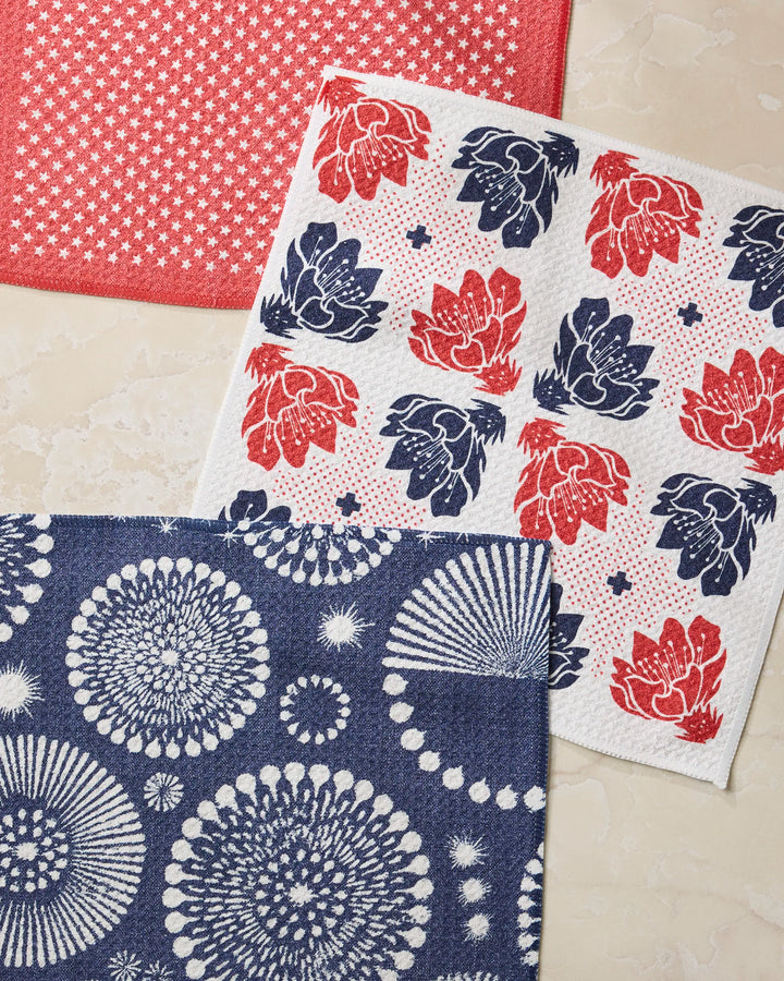 The Geometry Talon kitchen towels set includes three absorbent dishcloths: one red with white stars, one white with red and blue florals, and one navy blue with bursts, all showcased on a marble surface.