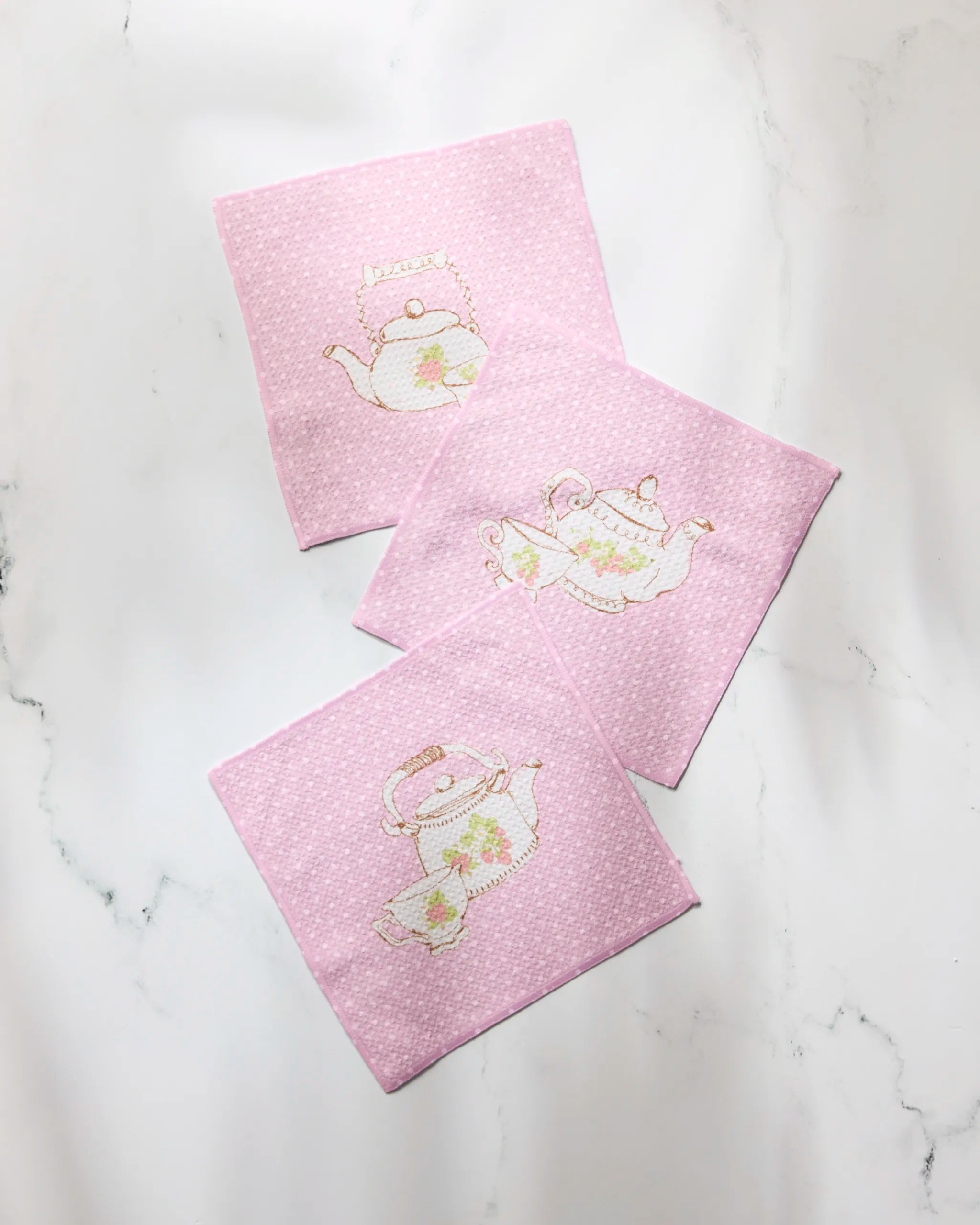 Three "Tea Time" coasters by Geometry, made from recycled pink dish towels with light polka dots and teapot embroidery, are displayed on a white marble surface.
