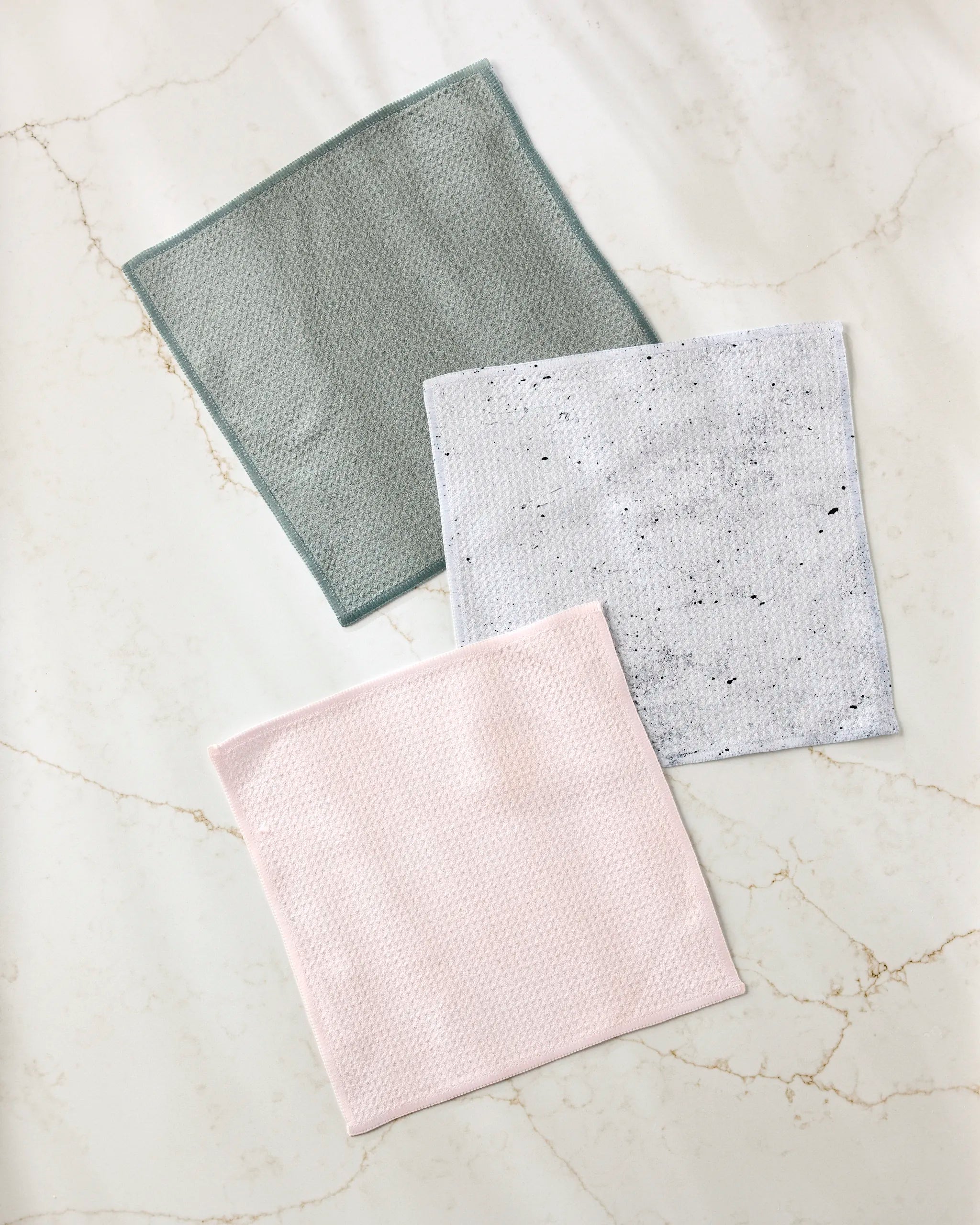 The Wright Set by Kailee Wright features three super-absorbent square dishcloths in light grey, pale pink, and white with black speckles, displayed on a white marble surface.