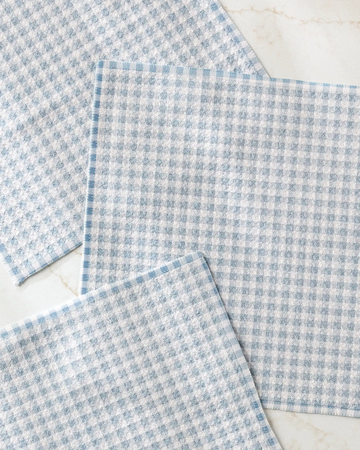 Three Tidepool Check cloths by Jenny Keller, featuring a light blue and white checkered textured pattern and made from recycled materials, are laid out overlapping at the corners on a white marble surface.