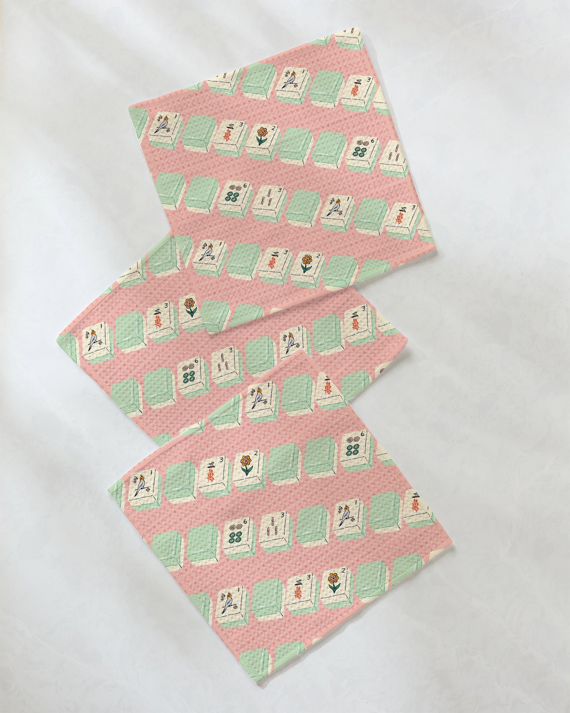 Three pastel pink and green cloths from Geometry’s Tile Parade super-absorbent dishcloth set, featuring a repeating Mahjong tile pattern, are arranged diagonally on a white surface.