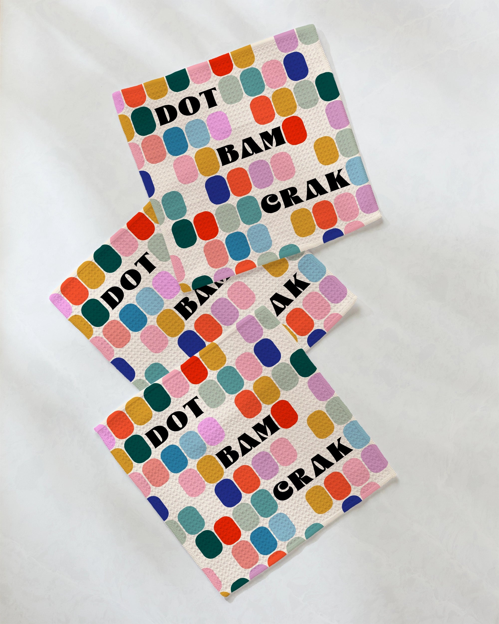 The Geometry "Tile Talk" dishcloth set features three vibrant napkins with multicolored dots and bold black lettering—“DOT,” “BAM,” and “CRAK”—arranged on a crisp white background, making it the perfect playful addition to your kitchen décor.