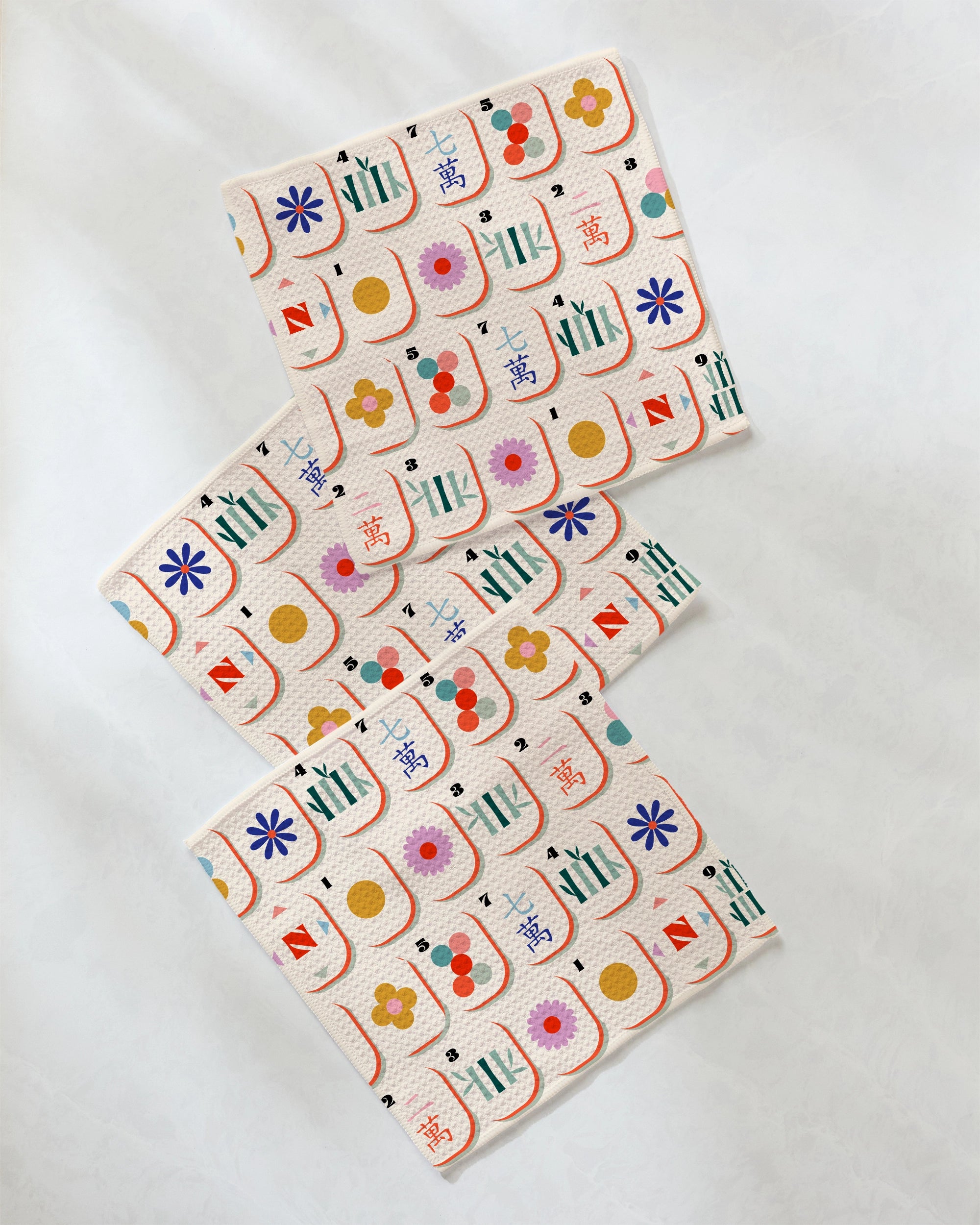 The "Tile Toss" dishcloth set by Geometry features three vibrant cloths adorned with colorful floral, circular, and geometric prints, arranged in a zigzag layout on a white background. Crafted from recycled materials, this eco-friendly set adds a lively touch to your kitchen.