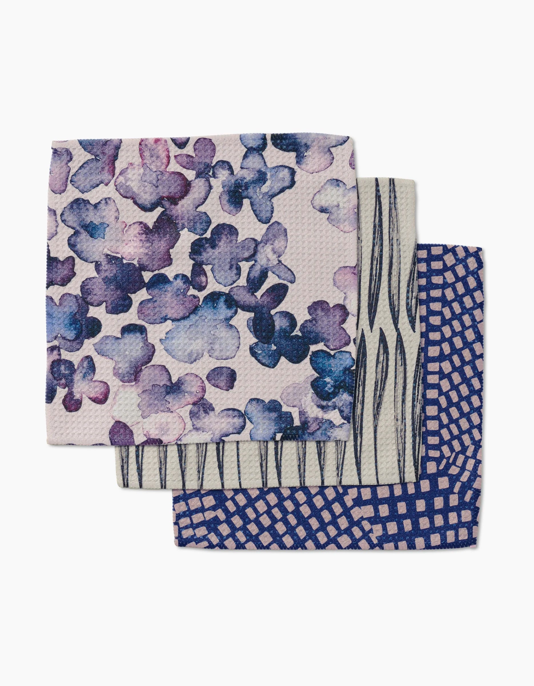 Three stacked GEOMETRY dishcloths (Tiny Petals): top has purple/blue watercolor flowers, middle features black vertical lines on white, and bottom shows a blue/pink geometric square design—all crafted with Geoweave fabric.