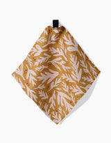 Trail Dusting Gold by Twig + Spark is a square tan cloth with a white botanical leaf pattern, made from recycled materials and hanging from a black loop against a white background.