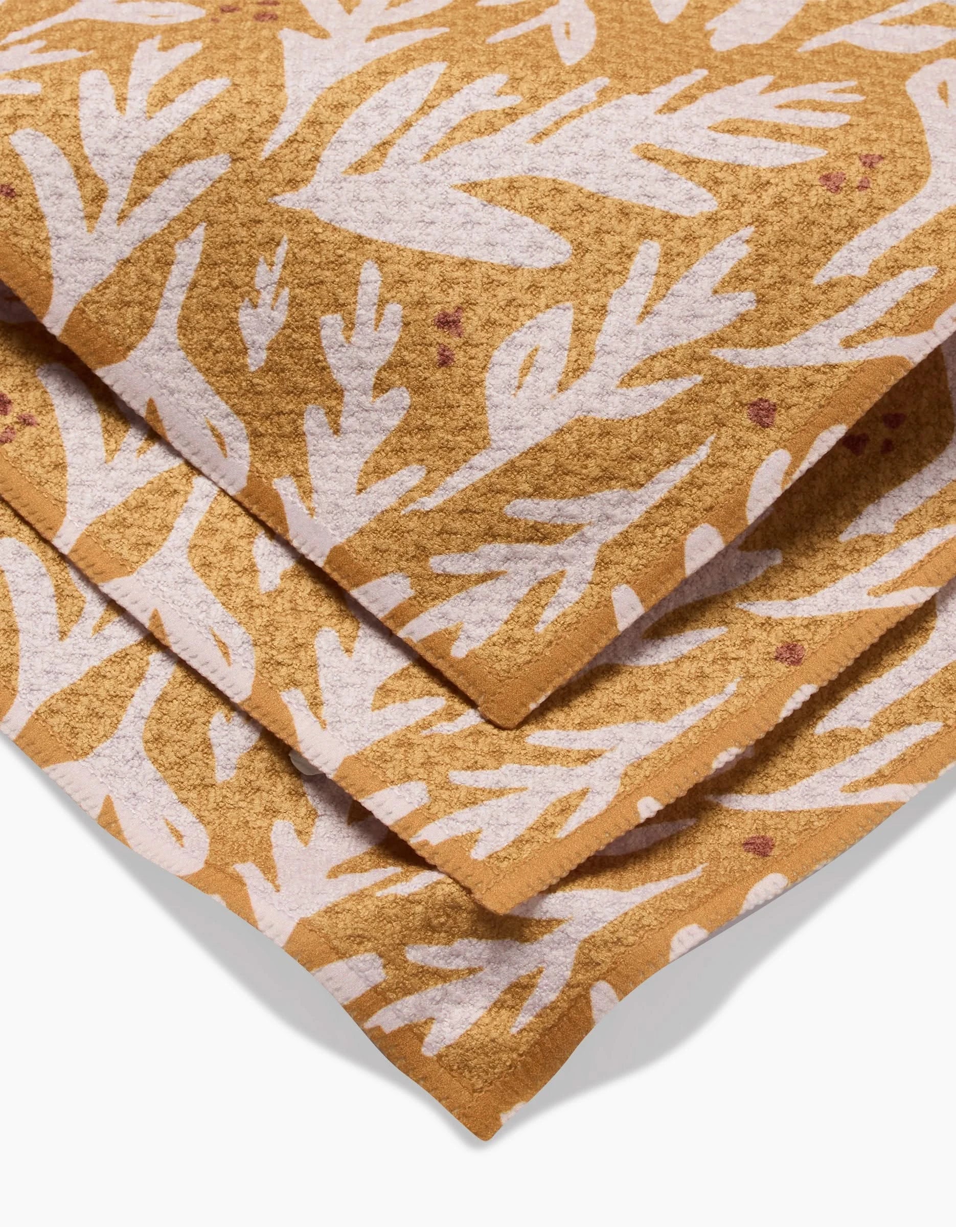 A close-up of folded Trail Dusting Gold towels by Twig + Spark, made from recycled materials, featuring a white leaf or coral pattern with small red dots. The overlapping towels highlight their textured fabric.
