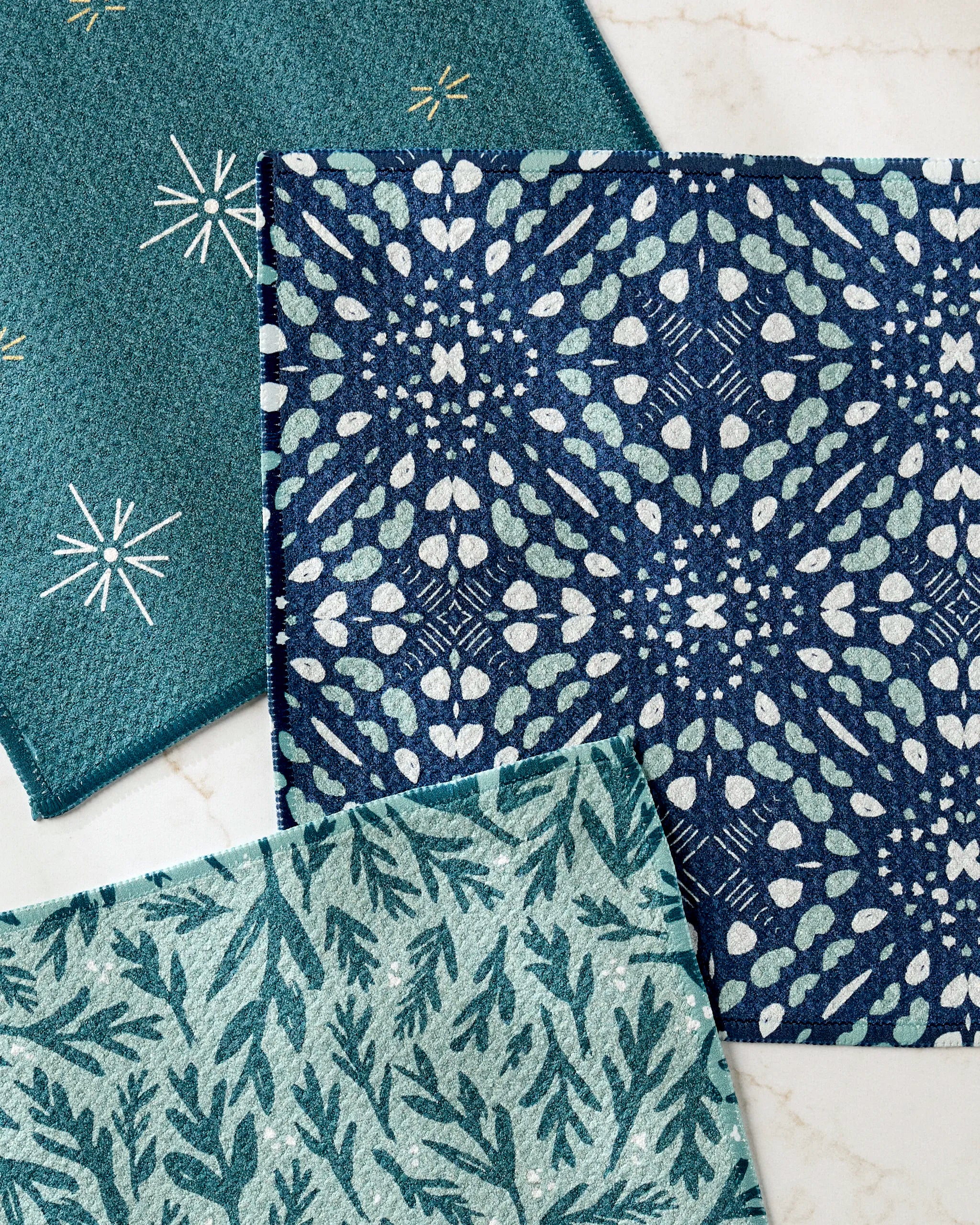 The Trail Dusting dishcloth set by Twig + Spark includes three super-absorbent towels in teal and blue with star, leafy, and botanical patterns, elegantly showcased on a marble surface.