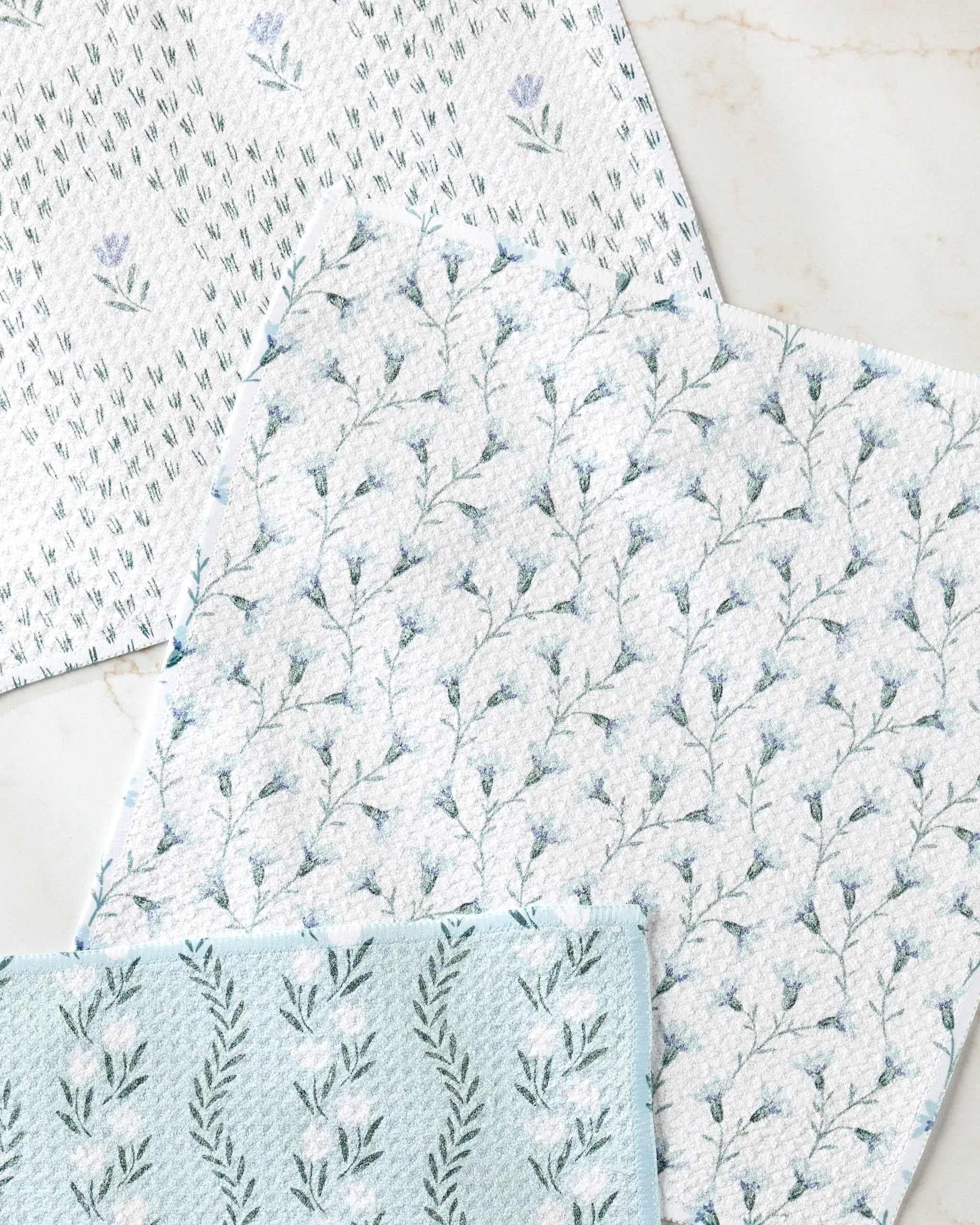 The Teri the Butter Trailing Farmhouse Florals set includes three absorbent dishtowels, each with a unique blue and green floral pattern featuring small flowers and leafy branches, displayed on a light marble surface.
