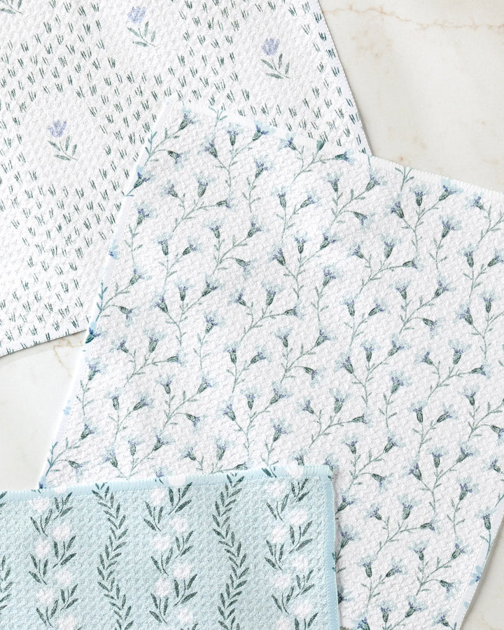 The Teri the Butter Trailing Farmhouse Florals set includes three absorbent dishtowels, each with a unique blue and green floral pattern featuring small flowers and leafy branches, displayed on a light marble surface.