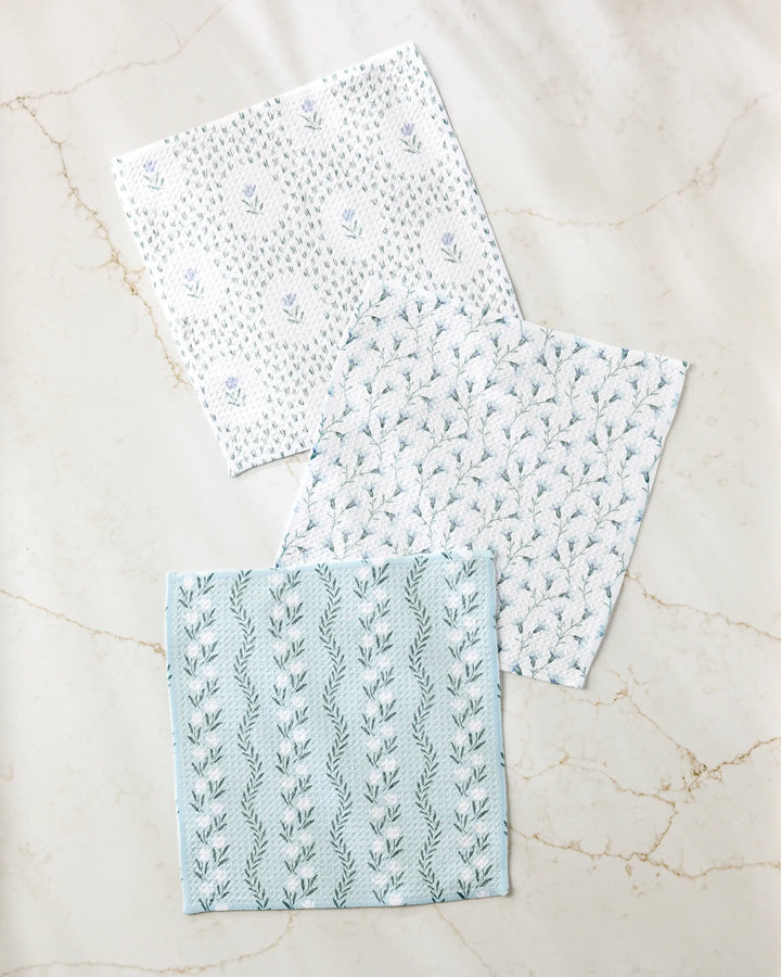 Three "Trailing Farmhouse Florals" napkins by Teri the Butter, featuring white and light blue botanical patterns and made from recycled materials, are arranged on a light marble surface.