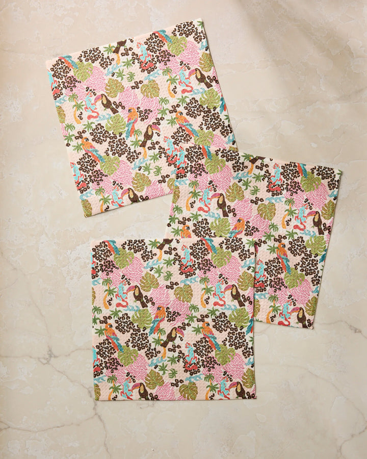 Three square napkins from Geometry's Tropi Hotel collection showcase a vibrant floral and leaf pattern in pink, green, brown, and blue, beautifully displayed on a light beige marble surface.